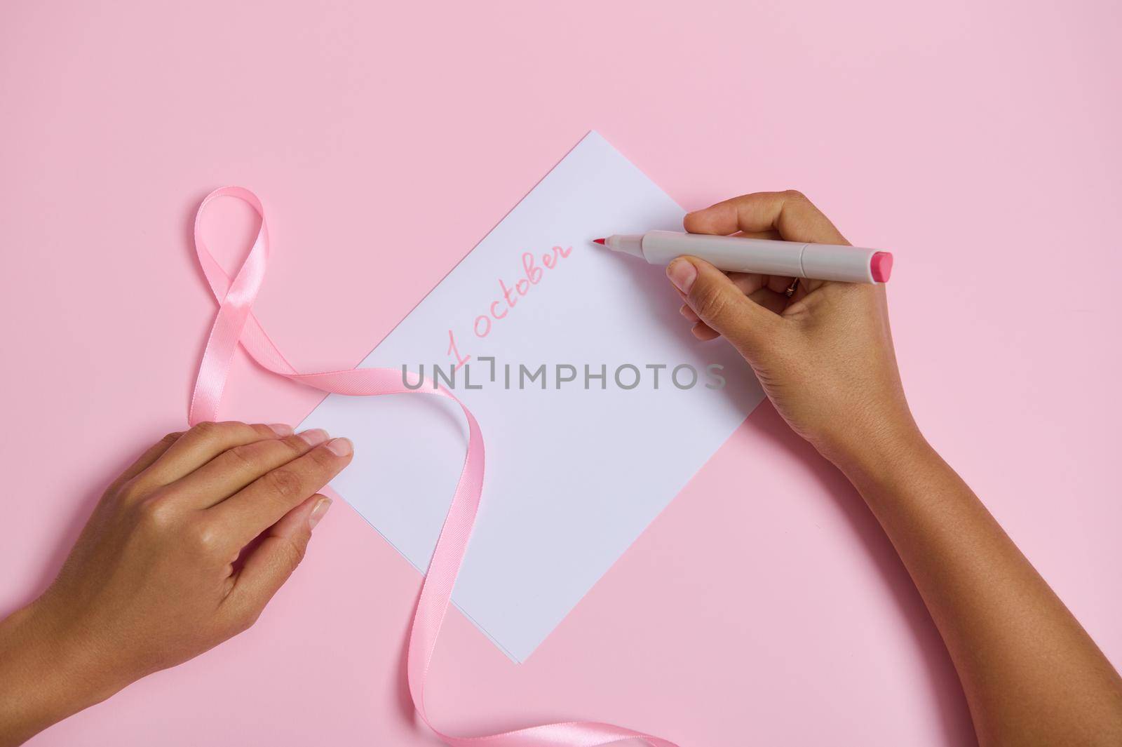 Flat lay of female hand holding felt pen, writes 1 OCTOBER on white blank sheet of paper, pink ribbon symbol of October Breast Cancer Awareness month, lying on pink background with space for text