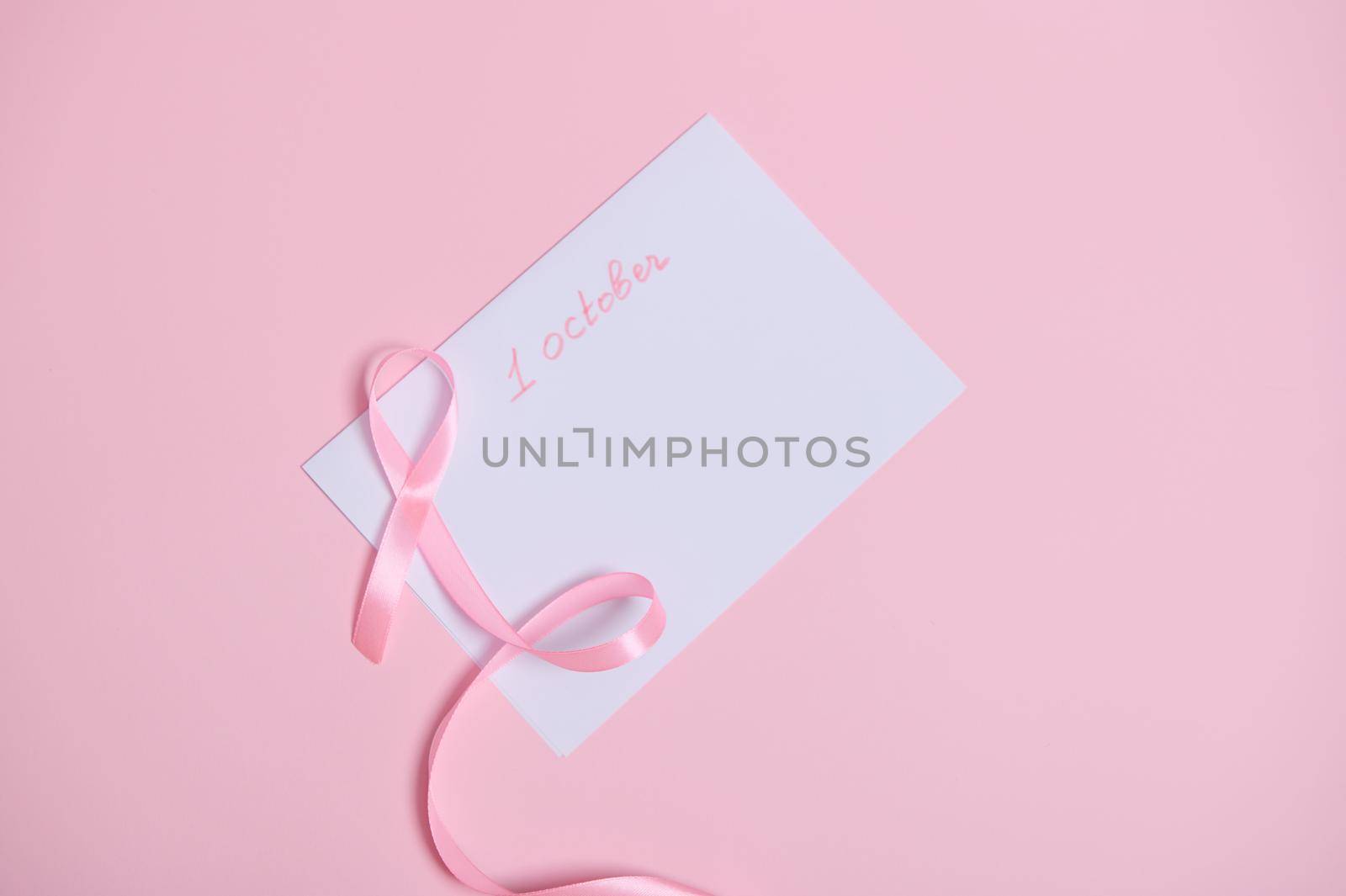 Flat lay of a pink ribbon with an endless end lying on a white paper sheet with lttering 1st October, International symbol of Breast Cancer Awareness Month. Woman's healthe care and medical concept by artgf