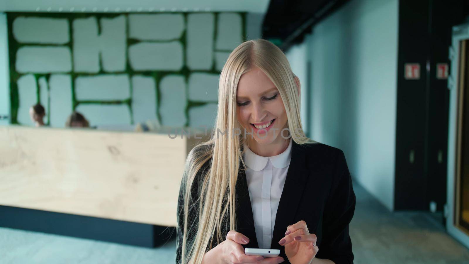 Cheerful young woman with phone in office. Pretty blond woman with long hair wearing elegant black suit and smiling charmingly at camera inside of modern office. by art24pro