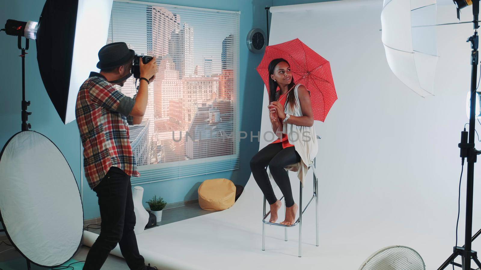 Stylish african model posing with red umbrella on bar high chair for fashion magazine photo shoot. Photographer taking photos on professional camera.