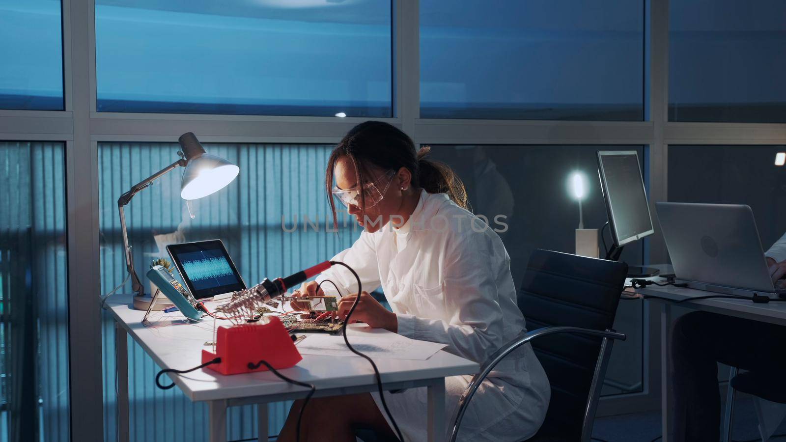 African american electronics specialist working with multimeter tester and other electronic devices in lab sitting at the table in protective glasses and white robe