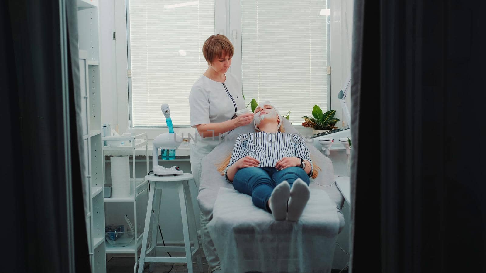 Cosmetologist applying cream mask on face of pretty woman at beauty spa salon. She making anti-aging treatments.