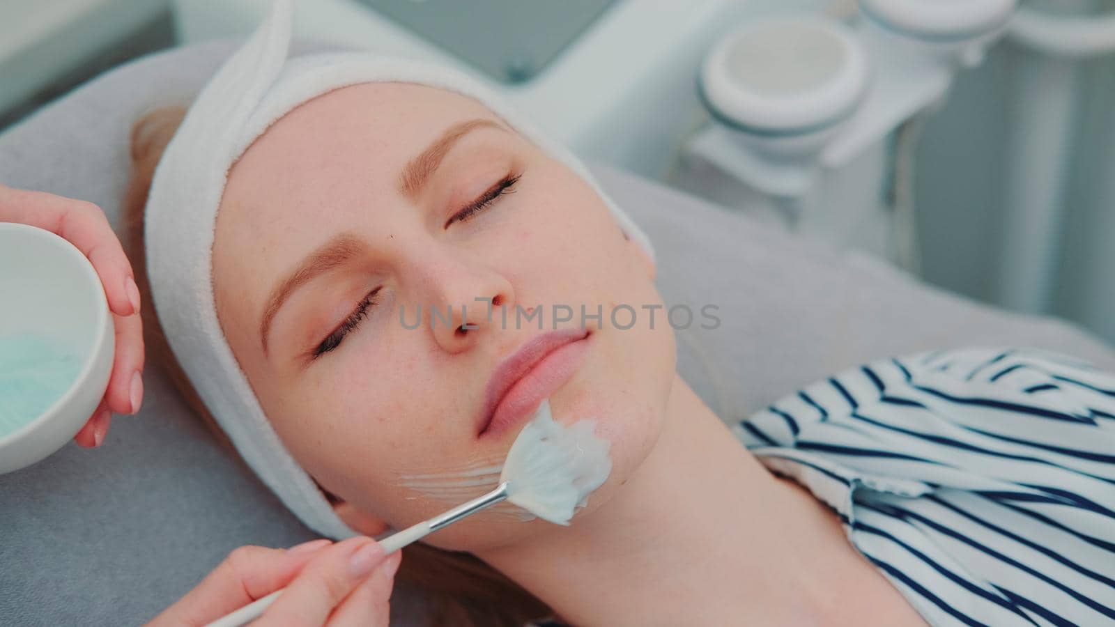 Cosmetician hands applying cream mask on young woman's face at beauty spa salon. Facial skin care treatments.