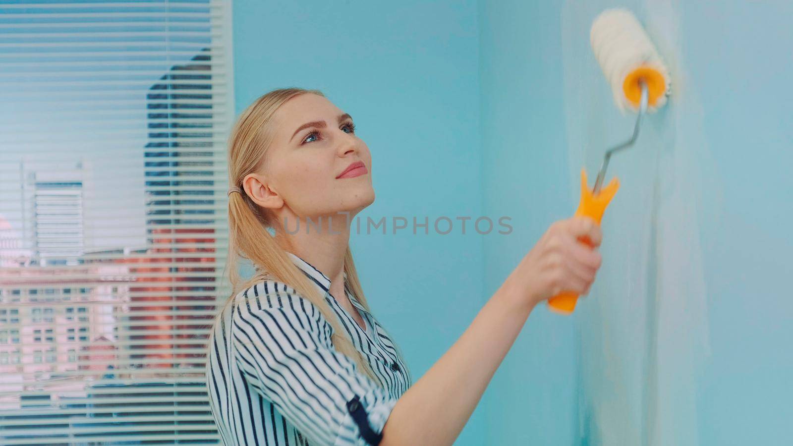 Close-up shot of woman builder at construction site with paint roller working, painting wall with white color in apartment. In the background there are skyscrapers at day.