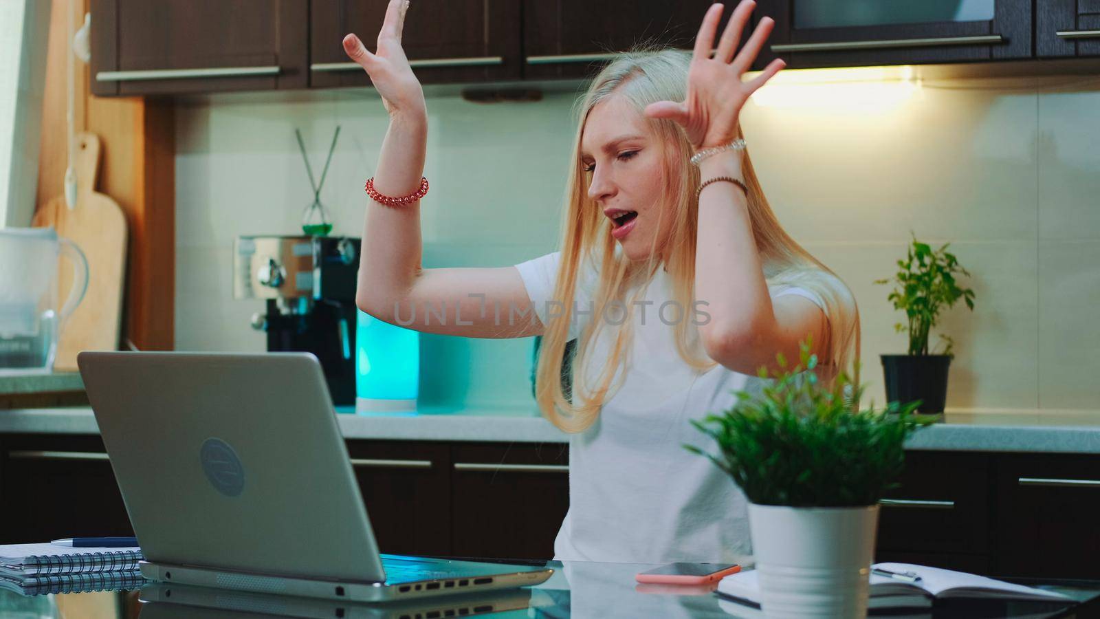 Blonde woman listening to the music and raising her arms in front of computer. She singing and sitting in the kitchen at home.