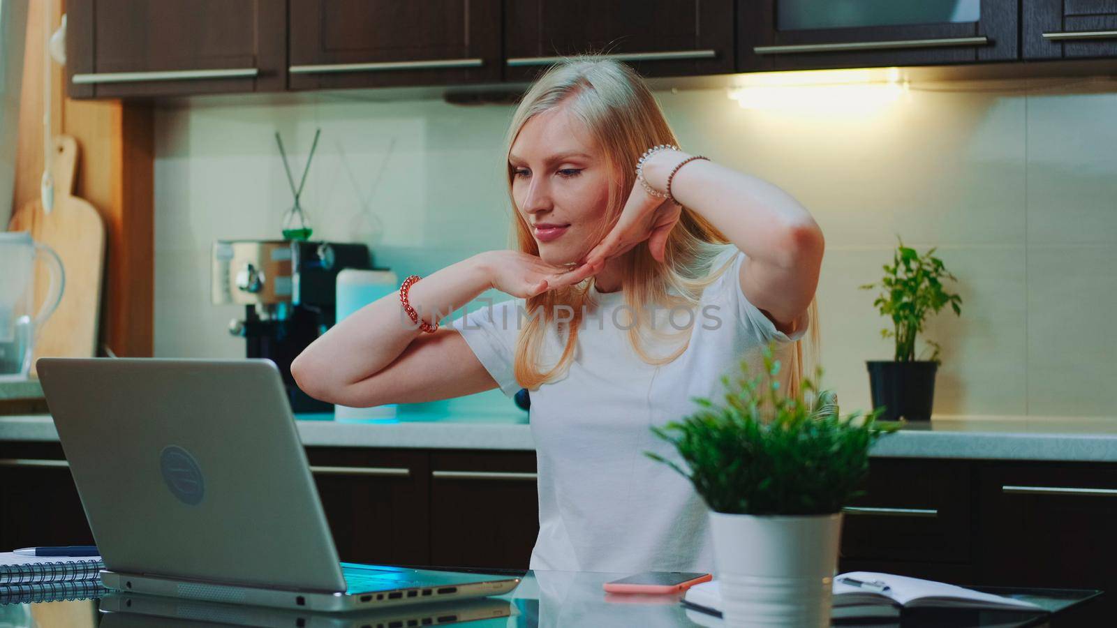 Blonde woman listening to the music and raising her arms in front of computer. She singing and sitting in the kitchen at home.