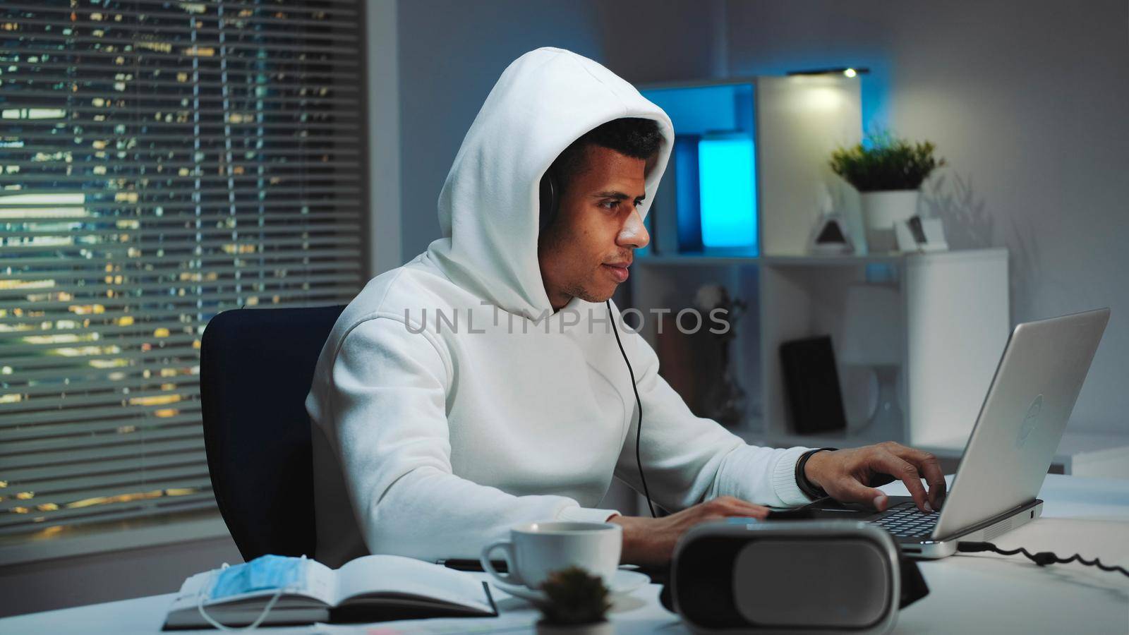 Black gamer in white hoodie and with headphones playing games on computer in the evening. He is passionate and smiled. Skyscrapers in the background.