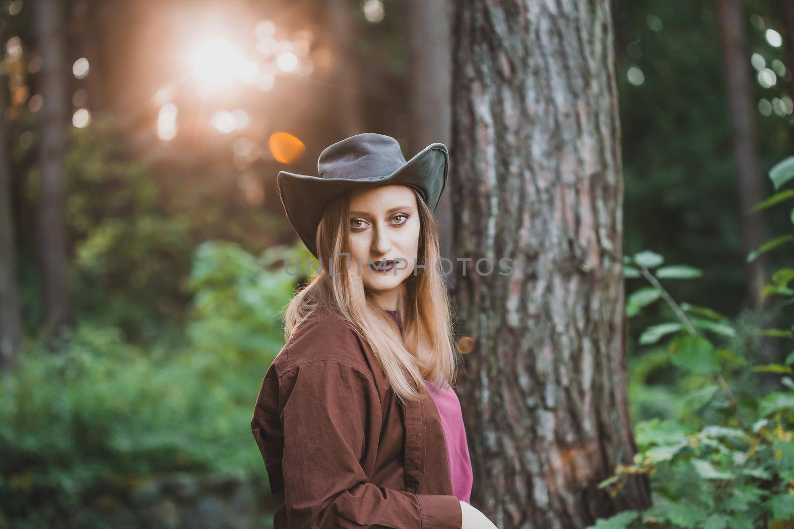 Country style fashion portrait of a blond woman wearing a leather cowboy hat and brown shirt standing in the forest. Dramatic heavy make up with dark lipstick. Halloween costume concept.