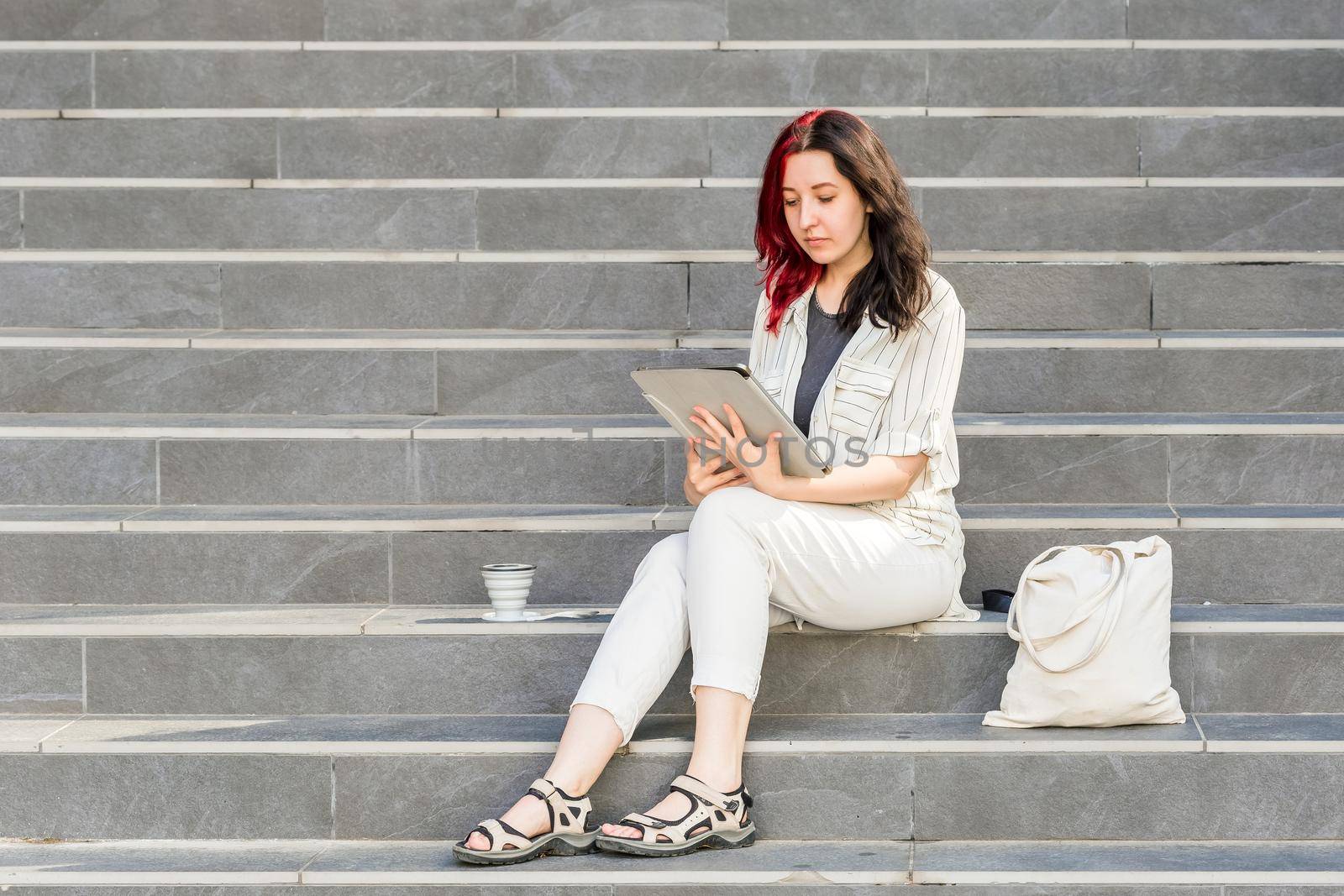 Beautiful White young business woman using Digital Tablet outdoors sitting on the stairs during her coffee break. Reusable coffee cup and linen shopping bag