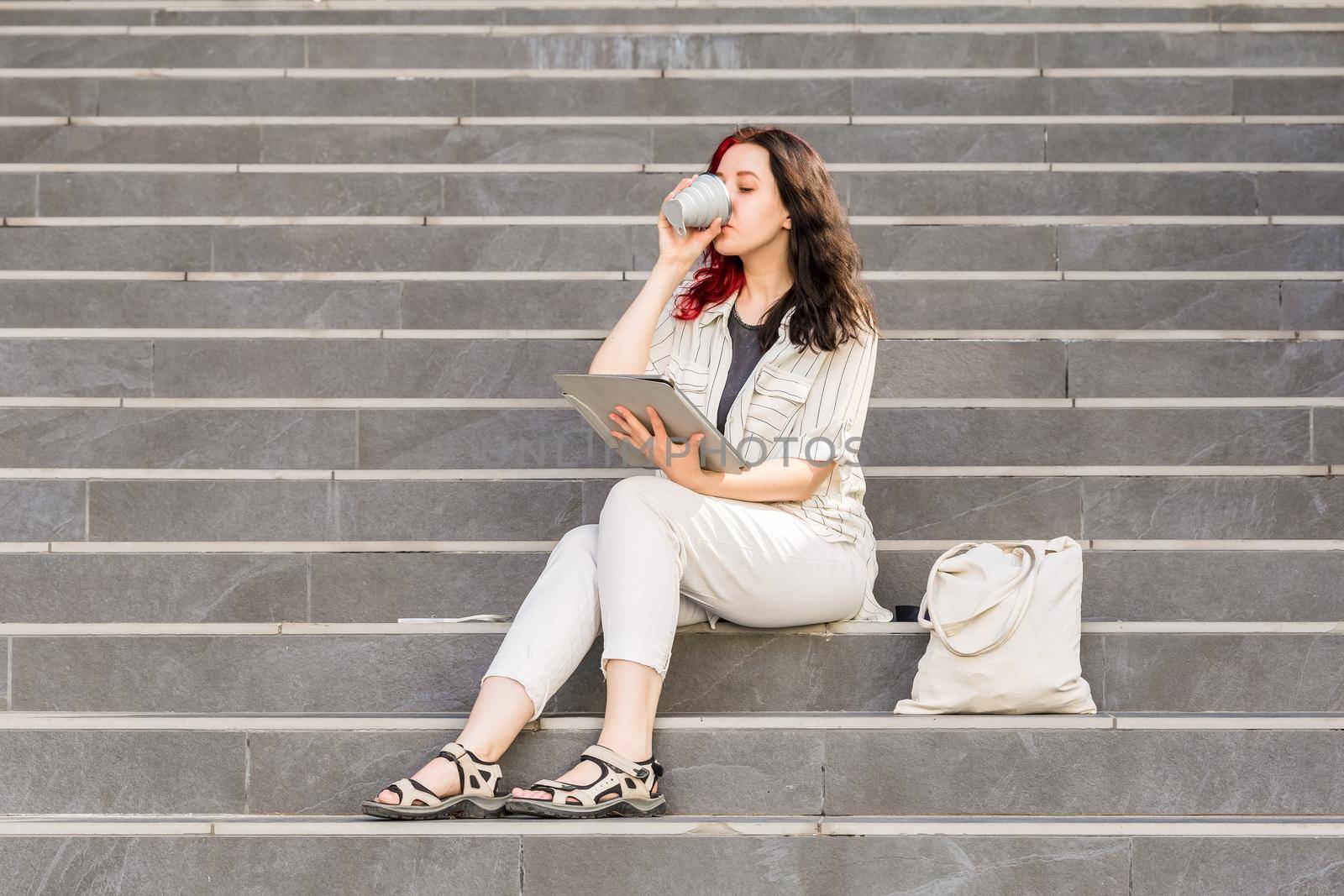 Beautiful White young business woman holding Digital Tablet outdoors sitting on the stairs during her coffee break. Drinking coffee from reusable collapsible cup, Sustainable lifestyle concept