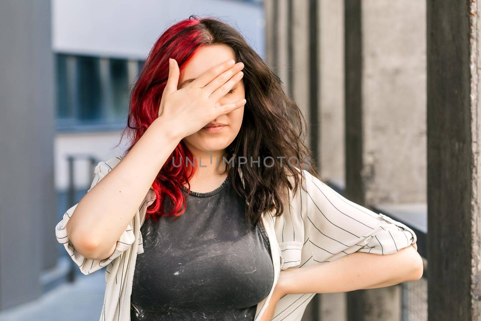 Stylish woman with red and black hair hiding eyes under hand while feeling ashamed or disappointed. Face palm gesture. Human facial expressions and emotions