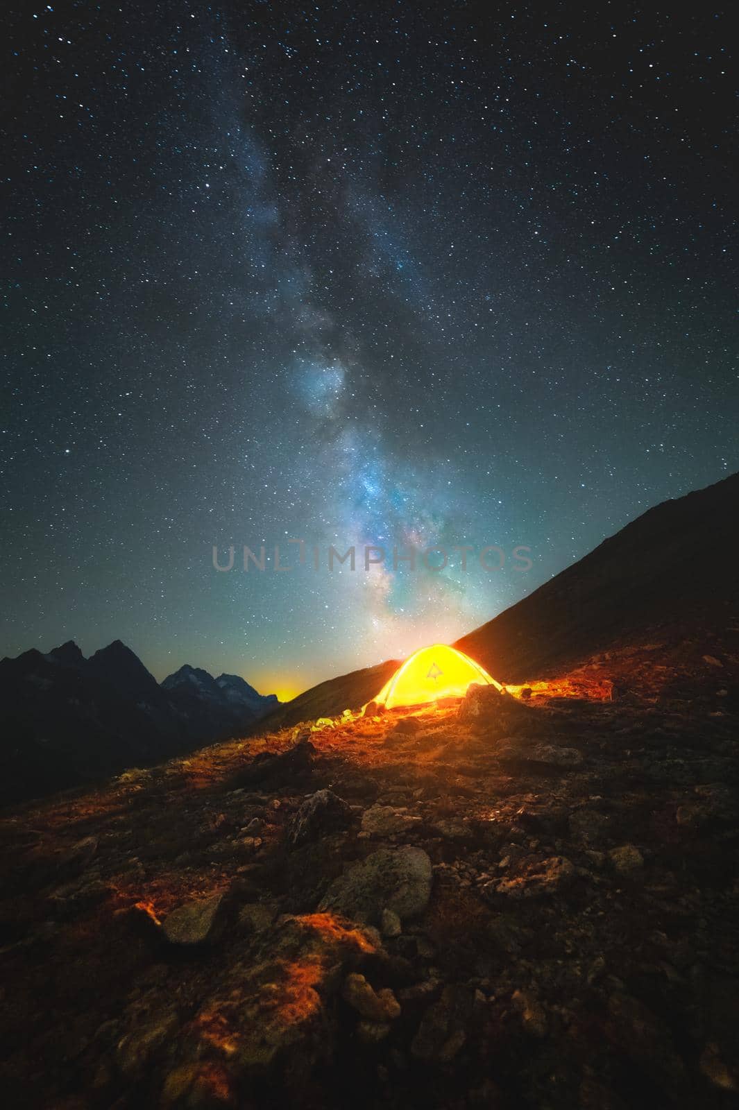 A vertical photo of the starry sky high in the mountains. The saturated milky way points to a luminous tent high in the mountains against the backdrop of a mountainous horizon. Astrophotography by yanik88