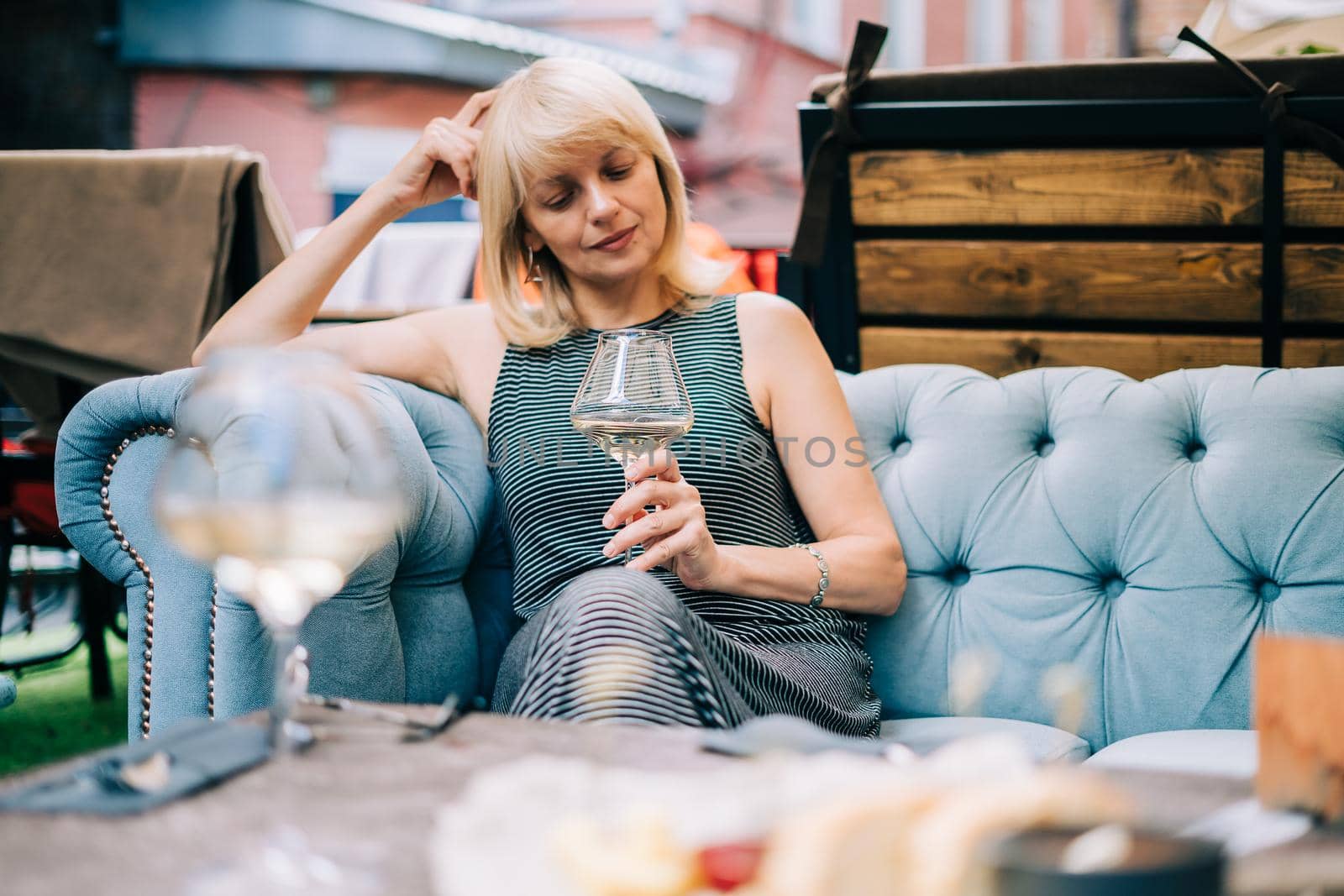 Mature adult woman sitting in bar outdoors with wine glasses and blurry restaurant background scene, drinking white wine and eating cheese. Summer sunny day on patio outdoors.