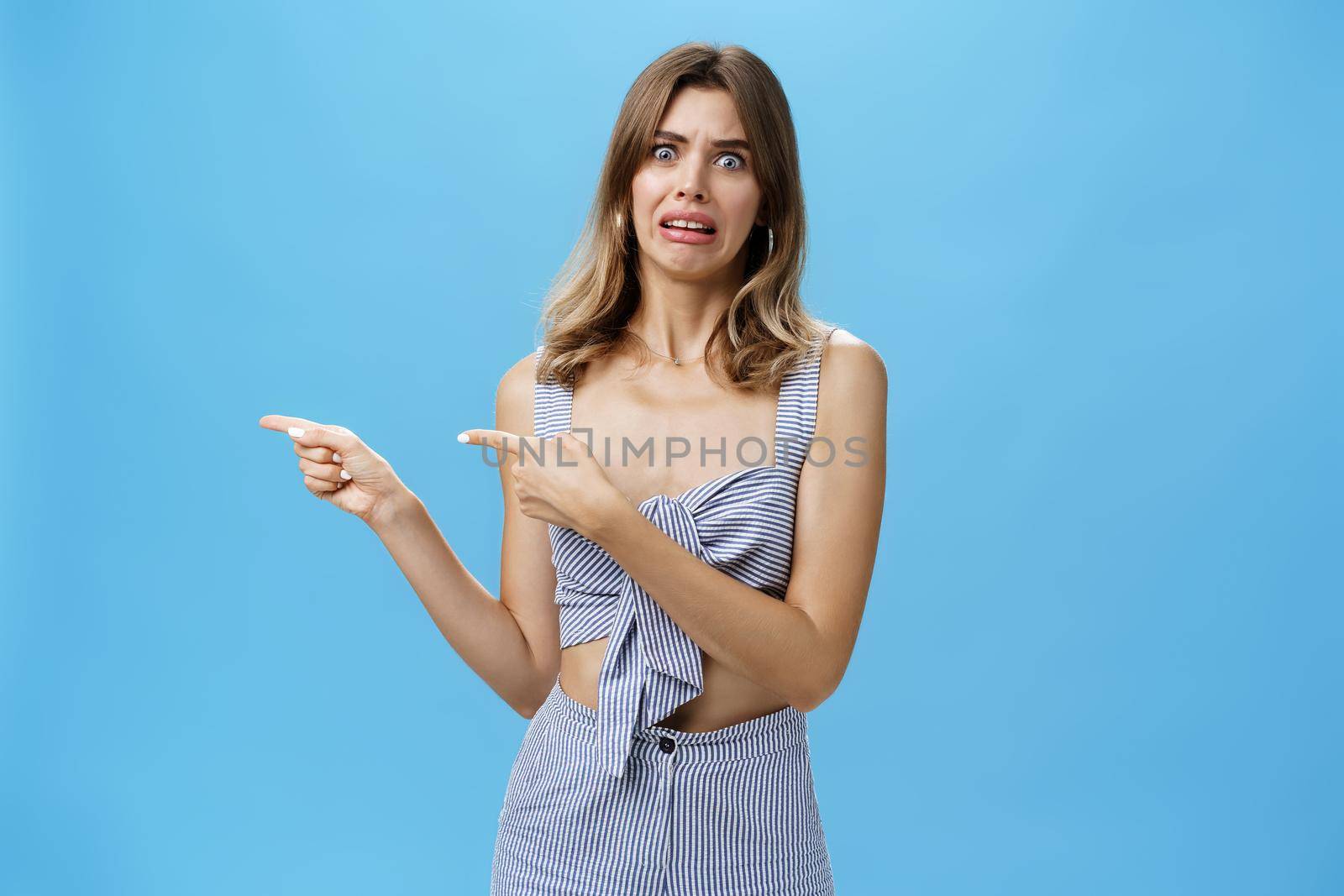 Studio shot of awkward displeased woman pointing left with index fingers making disgusted and dissatisfied expression grimacing from aversion seeing something creepy and awful over blue background. Lifestyle.