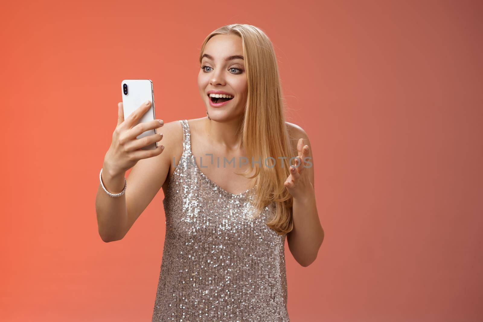Charming elegant nice blond girl in silver dress talking video call speaking looking smartphone display amused surprised smiling happily have conversation sibling showing prom outfit by Benzoix