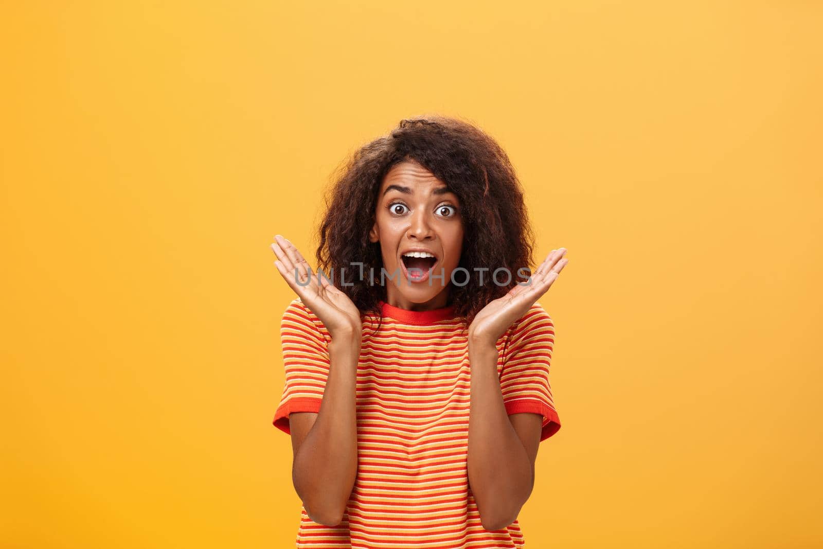 Waist-up shot of amazed joyful sociable dark-skinned female with afro hairstyle in striped t-shirt gesturing with palms raised near face gazing astonished and delighted at camera from surprise and joy. Lifestyle.