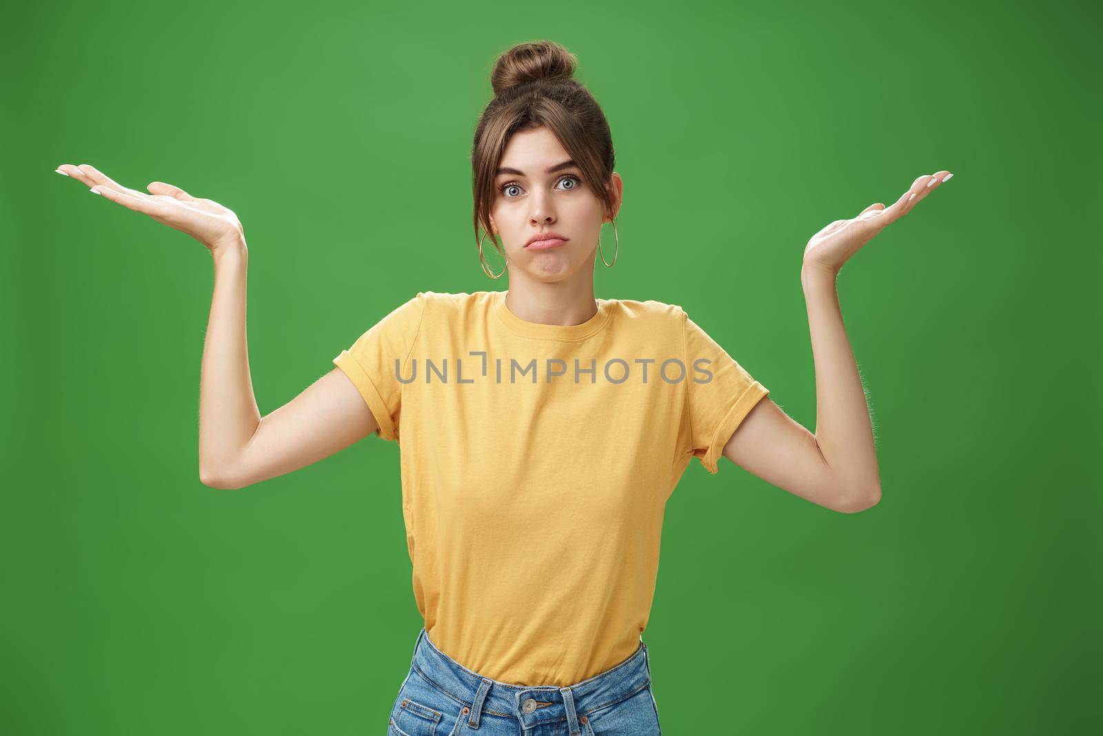 Girl do not know anything. Portrait of confused unsure cute woman with combed hair shrugging with raised hands pouting looking clueless at camera standing questioned against green background. Body language concept