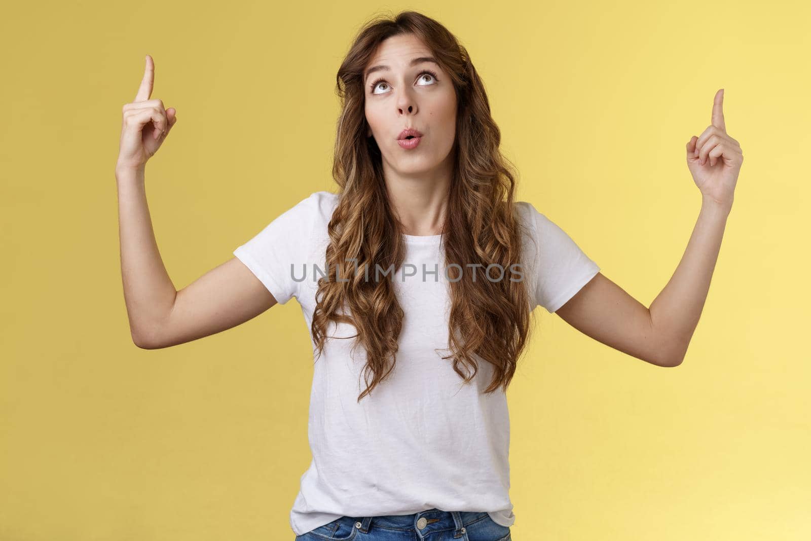 Impressed curious wondered attractive european woman long curly haircut look pointing index fingers up top copy space promo react astonished fascinated surprising event yellow background.