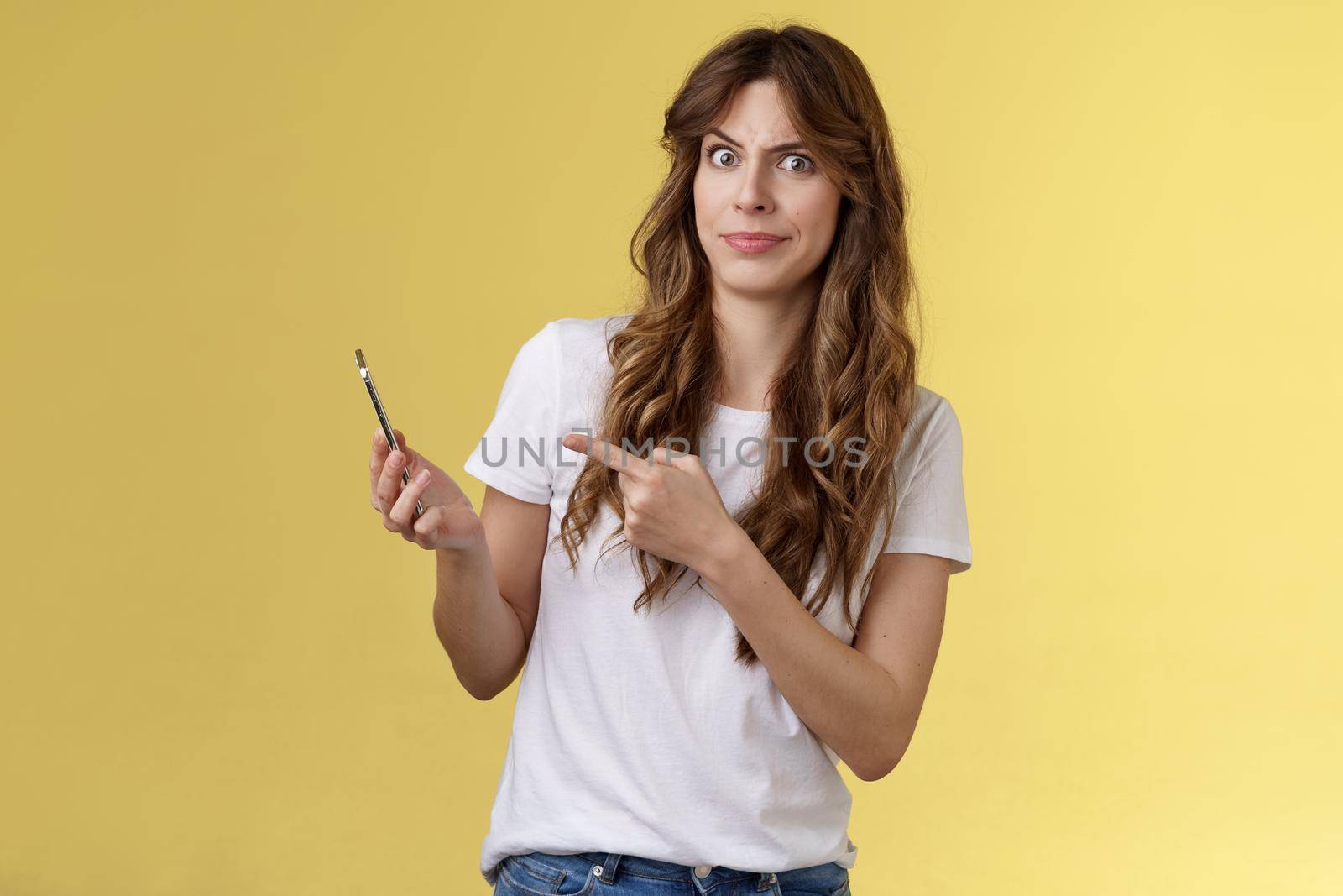 Girl answers strange weird call receive crazy upsetting message cringe doubtful displeased smirking dismay pointing suspicious smartphone stand yellow background intense frustrated.