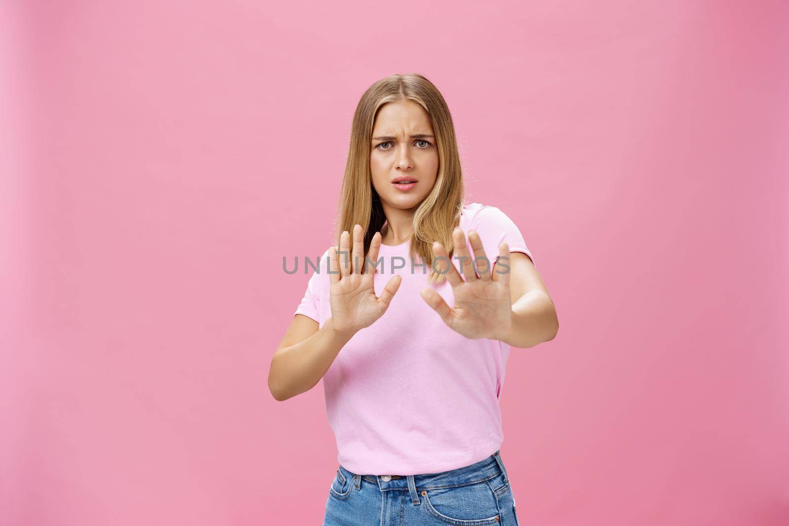 Stay away from me. Intense worried and displeased woman being victim of sexual harassment raising palms in defensive gesture making step backwards frowning asking stop, refusing over pink wall. Body language concept