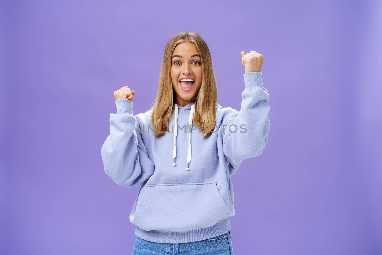 Cheerful happy and supportive young girlfriend with fair hair and tan in warm hoodide raising fists in cheer and triumph smiling saying yeah celebrating goal or success over purple background. Lifestyle.