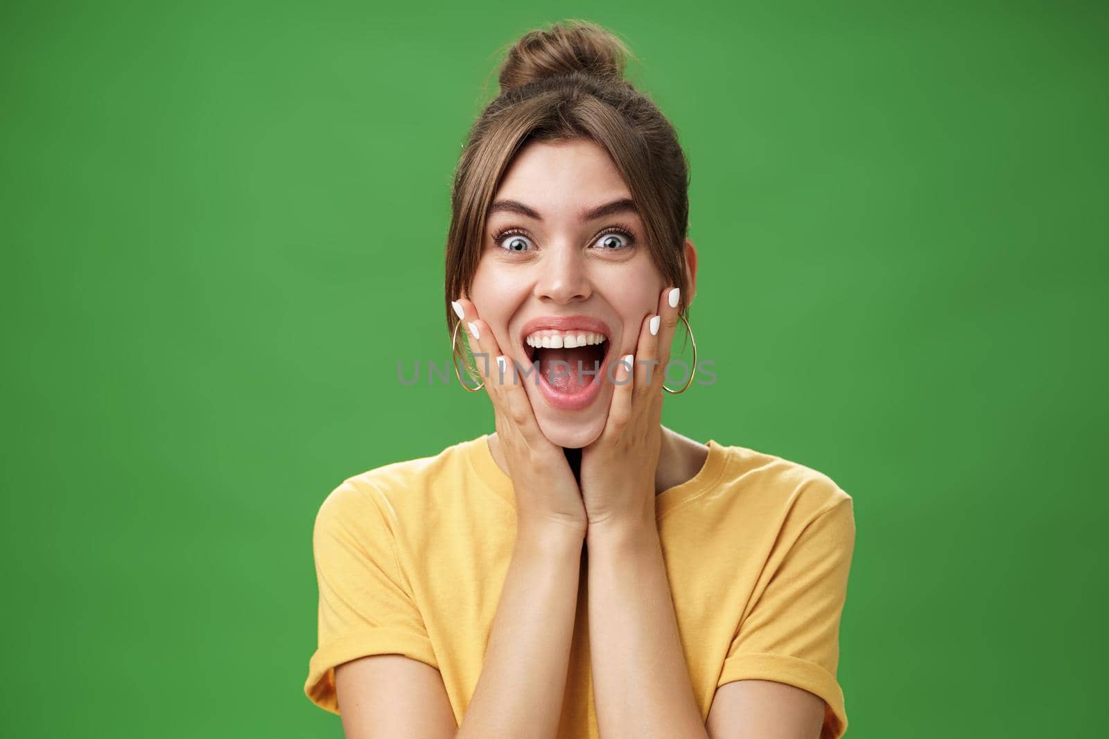 Portrait of happy delighted and surprised young feminine girl in yellow t-shirt pressing hands to cheeks from amazement and joy smiling broadly reacting to astonishing news over green background. Emotions concept