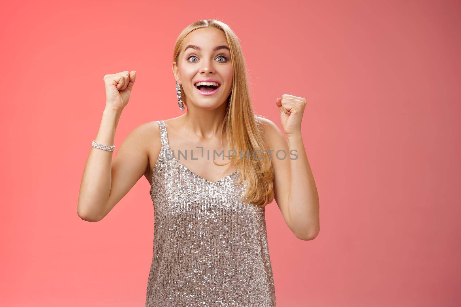 Lifestyle. Surprised happy celebrating blond young woman in silver trendy dress raising hands up yes victory gesture smiling broadly excited winning first place reach goal grinning thrilled triumphing.