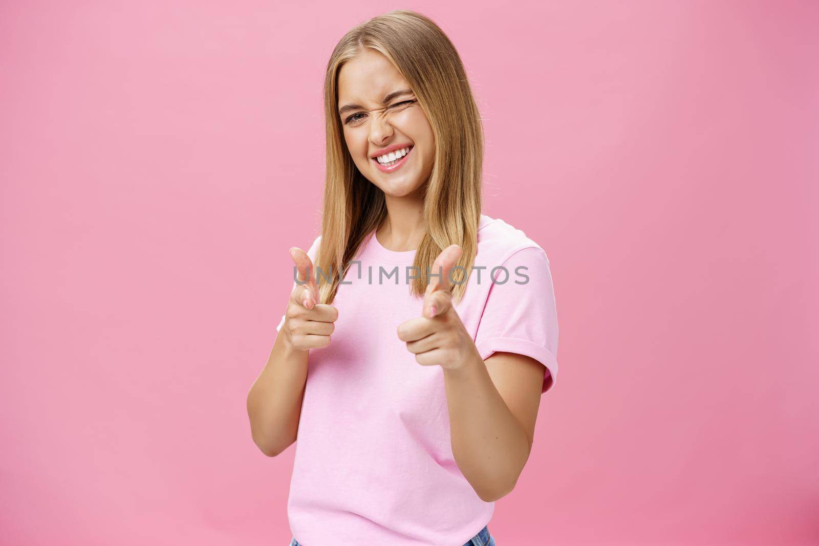 Woman expressing positive attitude towards camera pointing with fingers and winking joyfully smiling being uplifted, standing in good mood with optimistic gestures against pink background. Copy space