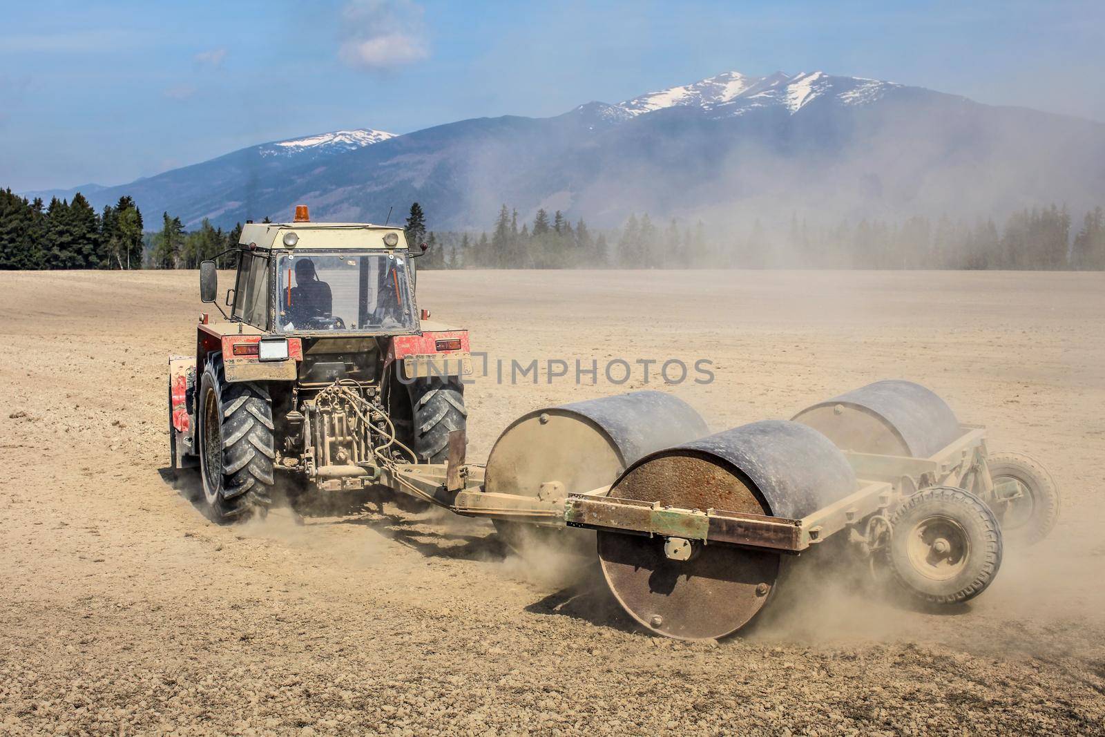 Tractor pulling heavy metal rollers on a dry field with mountains in background. Spring ground preparation.