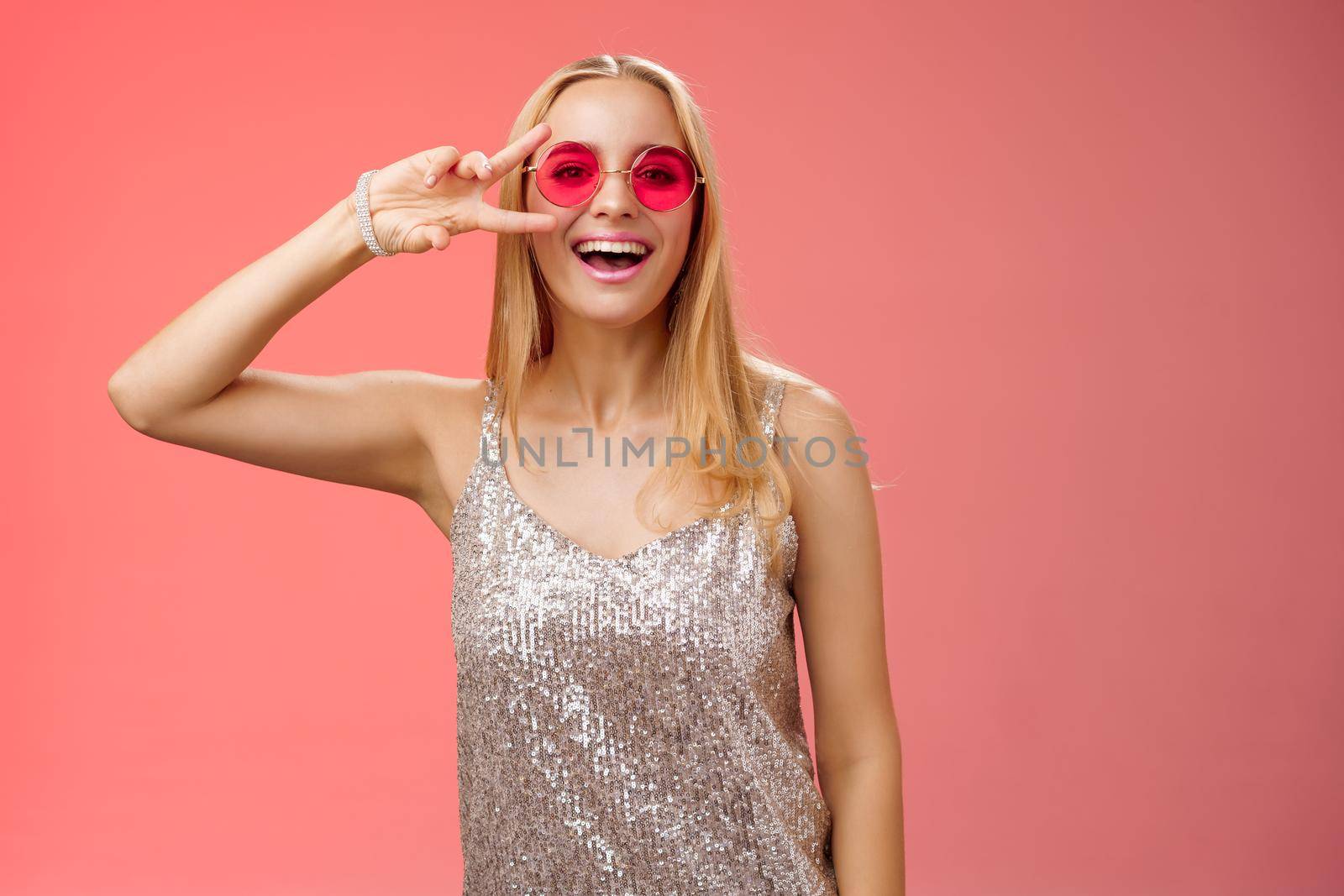 Energized friendly good-looking blond 25s woman in silver stylish dress sunglasses show peace victory gesture carefree smiling camera have fun enjoying cool party, standing red background.