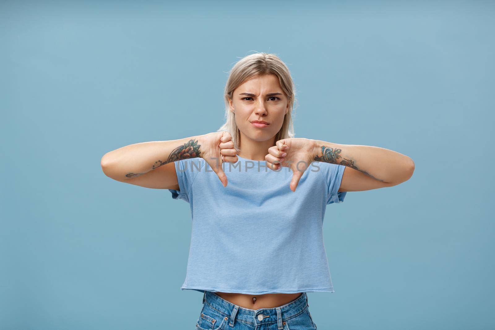 No way mate dislike. Portrait of dissatisfied bossy female tattoo artist with tattoos on arms frowning from displeasure showing thumbs down in disapproval standing over blue background. Copy space