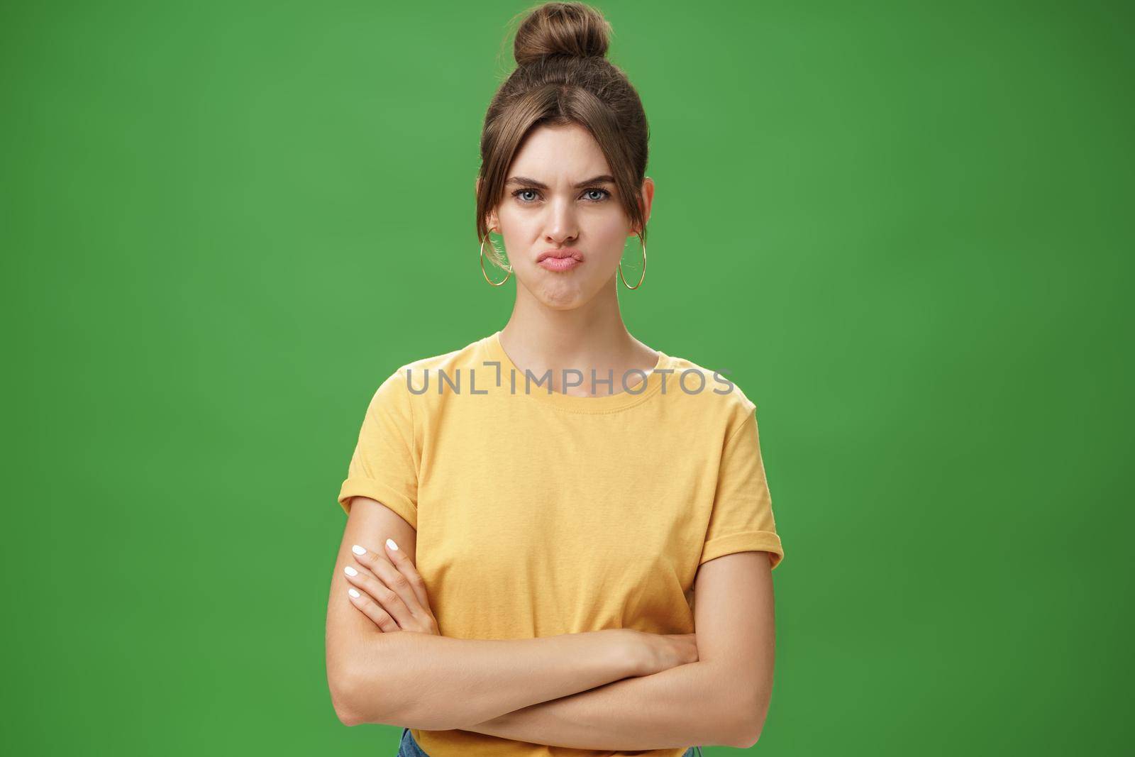 Girl not liking result of work looking displeased and serious at camera, frowning crossing hands against chest, pursing lips from disappointment, being offended over green background.