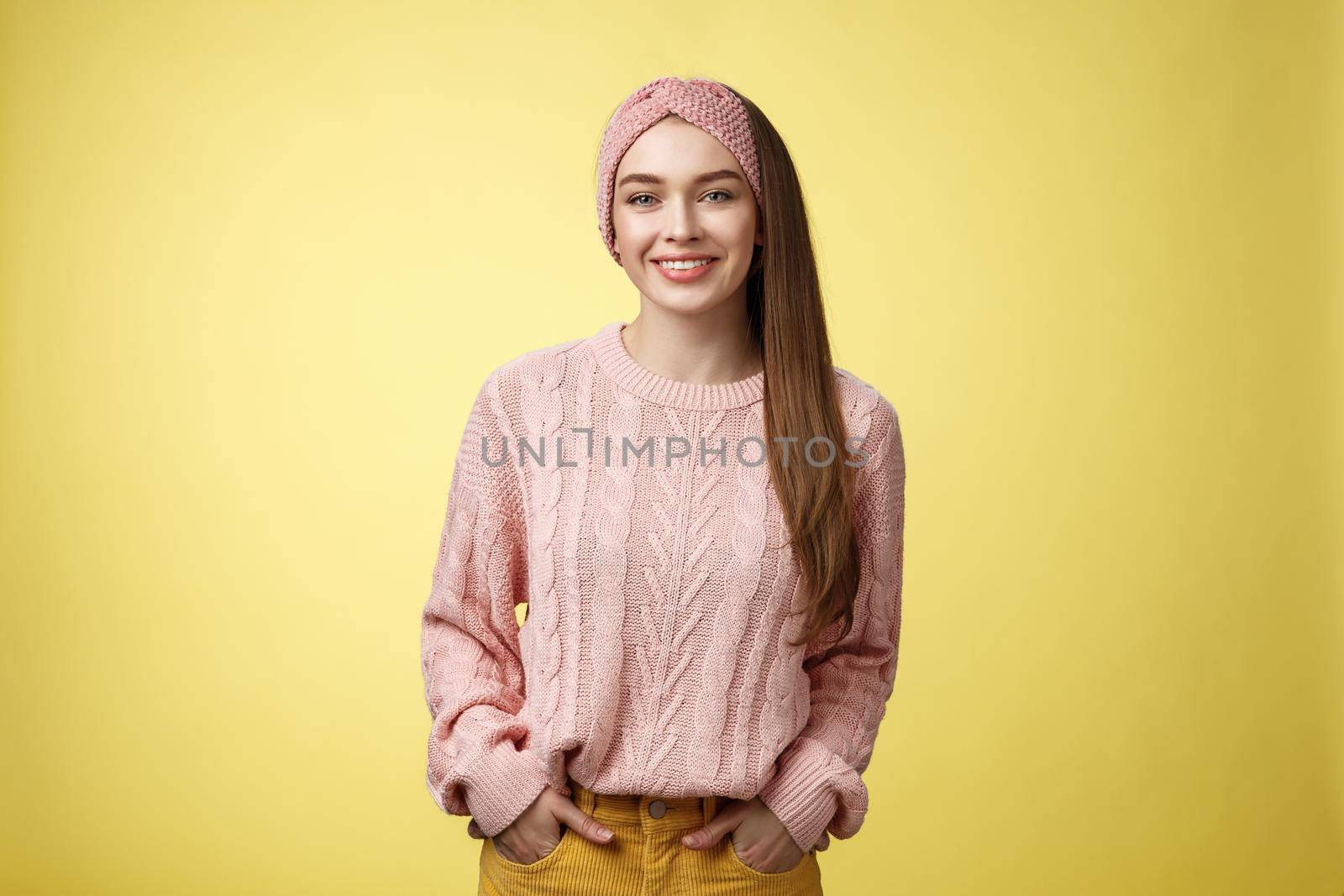 Charming friendly young girl in knitted sweater and headband holding hands in pockets, smiling, looking positive motivated achieving goals having great day, posing positive against yellow background by Benzoix