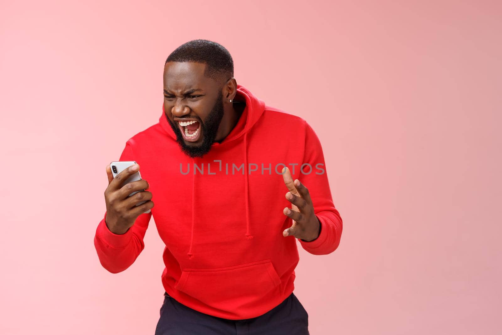 Angry furious african-american bearded guy lose control emotions shouting outraged annoyed smartphone display look phone insane stooping frowning rage, standing irritated pink background.