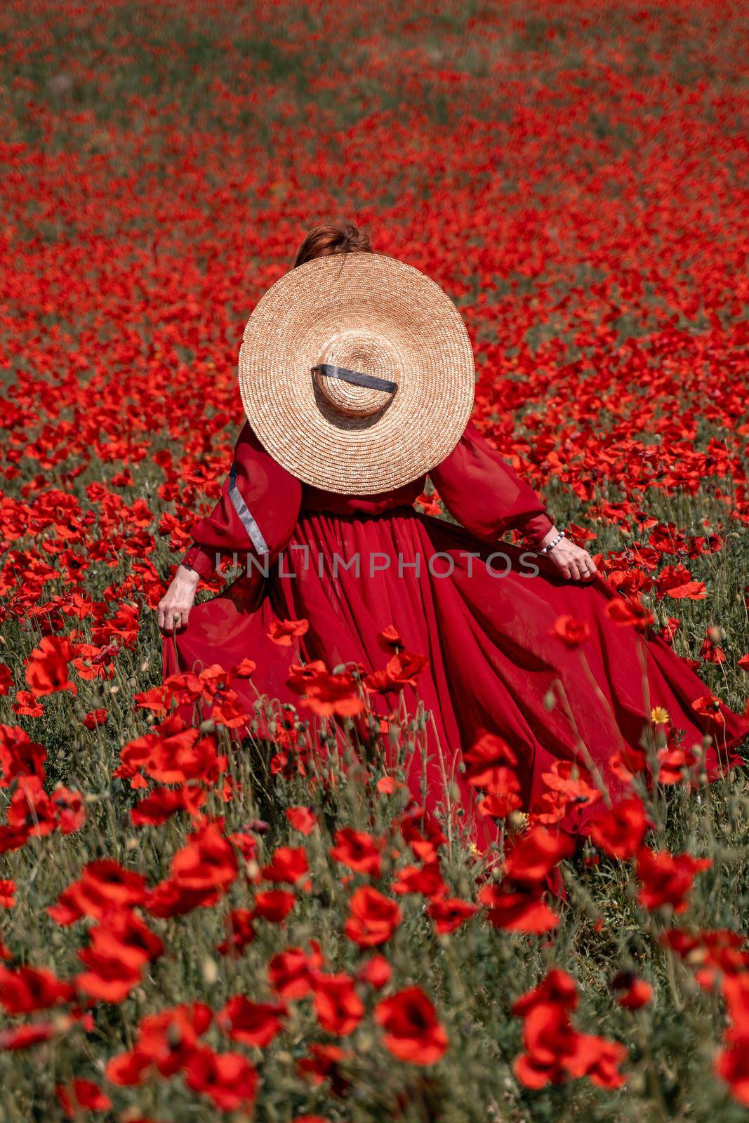 Young woman stands with her back in a long red dress and hat, posing on a large field of red poppies.