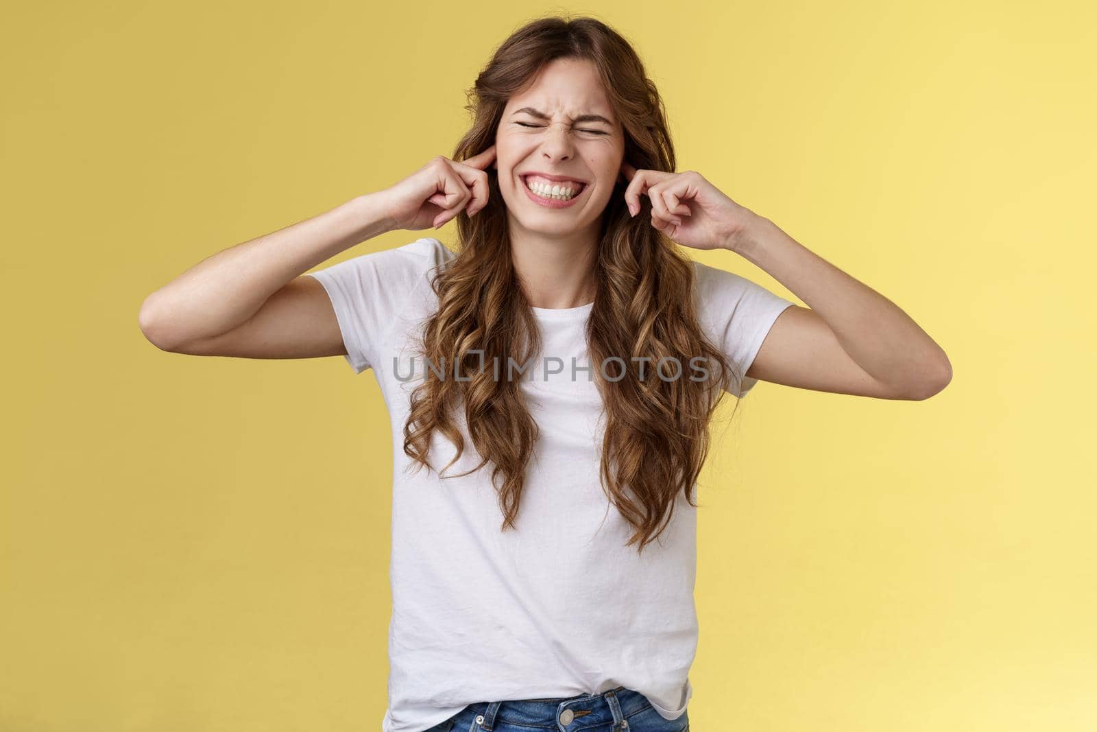 Turn this awful music off. Girl making wry face shut ears index fingers plugged earholes close eyes clench teeth displeased annoyed loud terrible noise irritated from neighbours yellow background by Benzoix