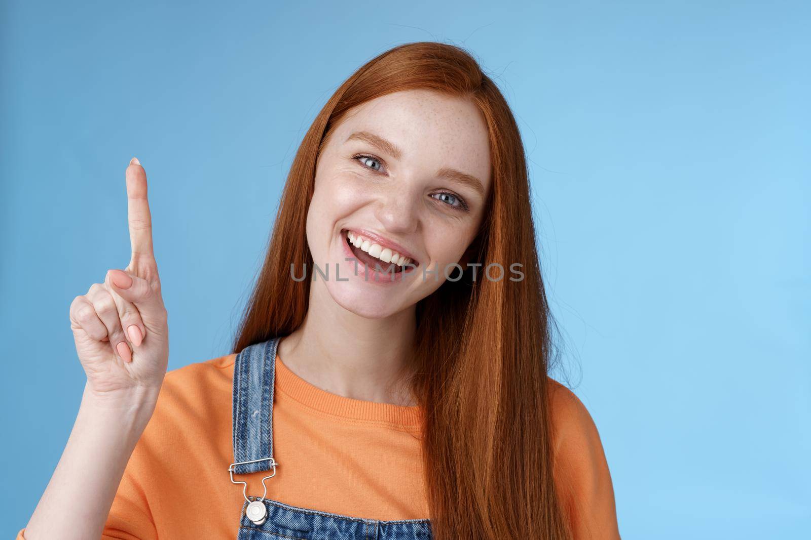 Lively active energized happy smiling hepful redhead female coworker showing you awesome promo pointing index finger up grinning introduce cool product recommend try out blue background by Benzoix