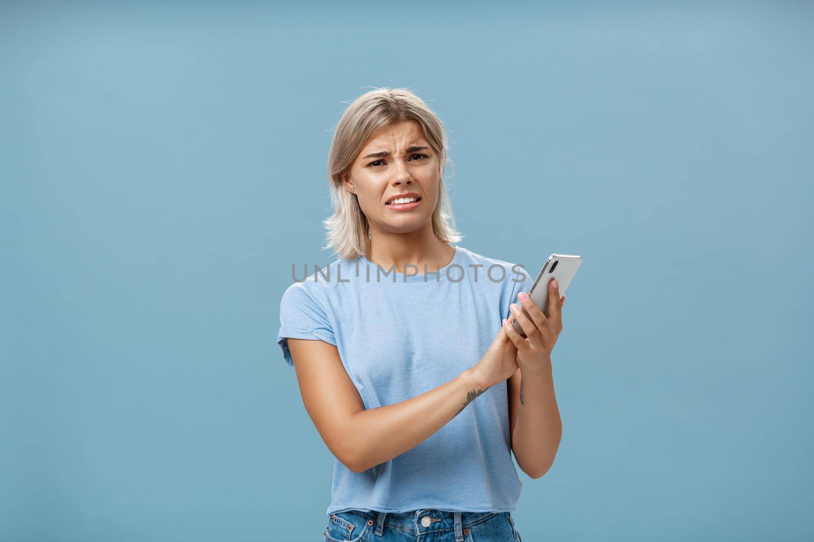 Woman cannot stand when mom shouts during phone call. Intense displeased young european blonde with tanned skin clenching teeth and frowning covering microphone of smartphone over blue wall.