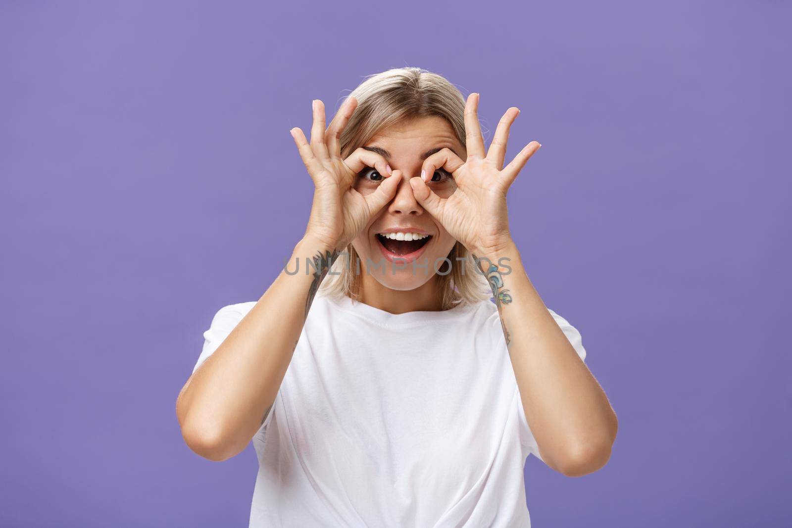 Looking at awesome discounts. Portrait of impressed amused and happy good-looking stylish young woman with tattoos on arms making circles with hands over eyes smiling with admiration at camera by Benzoix