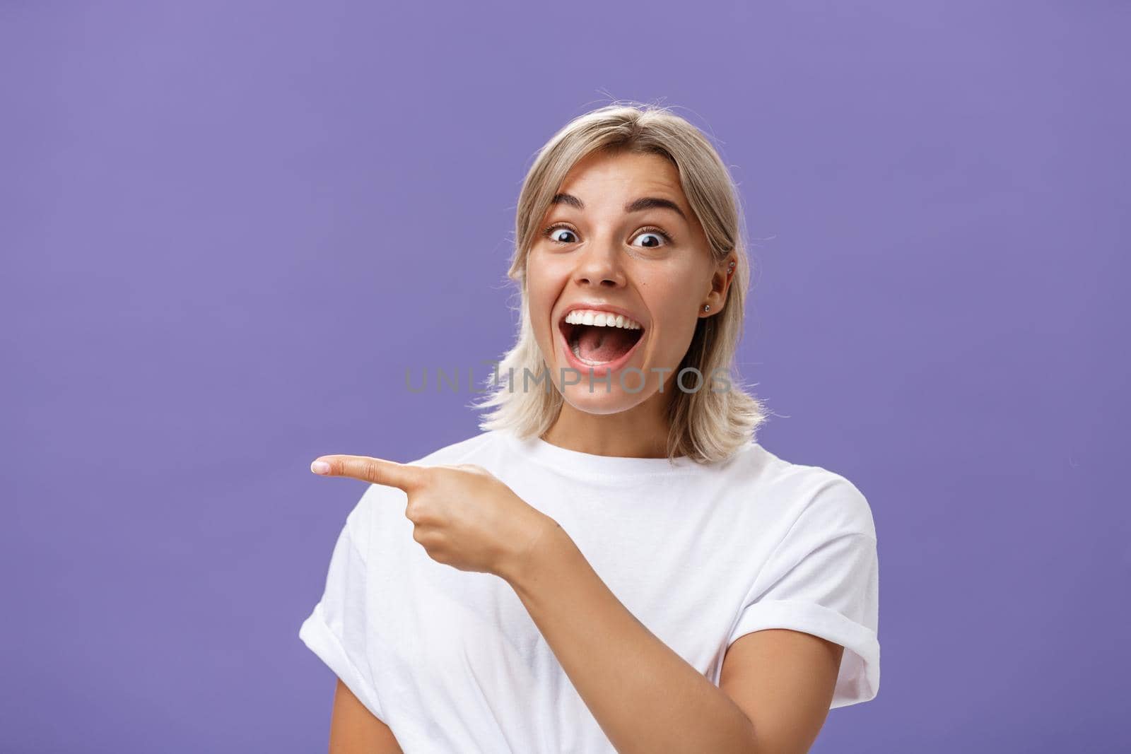 Waist-up shot of joyful thrilled attractive and emotive young blonde female with tanned skin in white casual t-shirt smiling and laughing happily pointing left with excitement over purple wall. Emotions concept