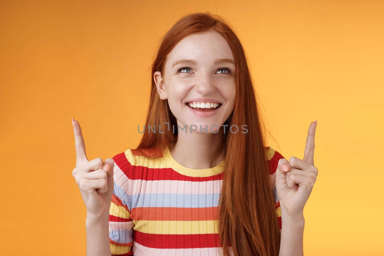 Happy cheerful redhead girl having fun amusement park laughing joyfully pointing look up index fingers upwards enjoy entertainment standing orange background amused grinning joyfully.