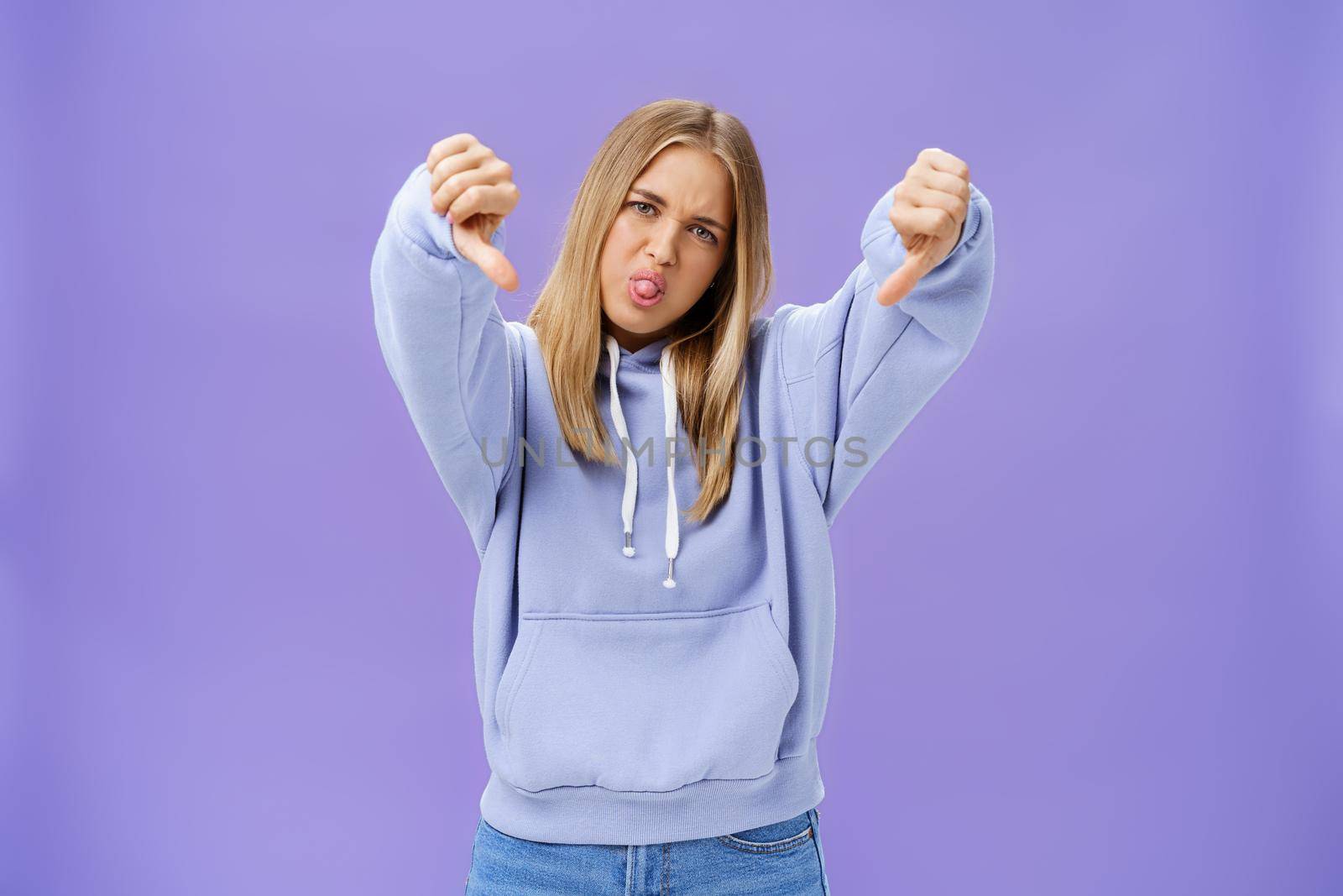 Indoor shot of moodie displeased and unimpressed cool modern female with blond hair and tanned skin tilting head sticking out tongue showing thumbs down in dissatisfaction posing against purple wall. Body language concept