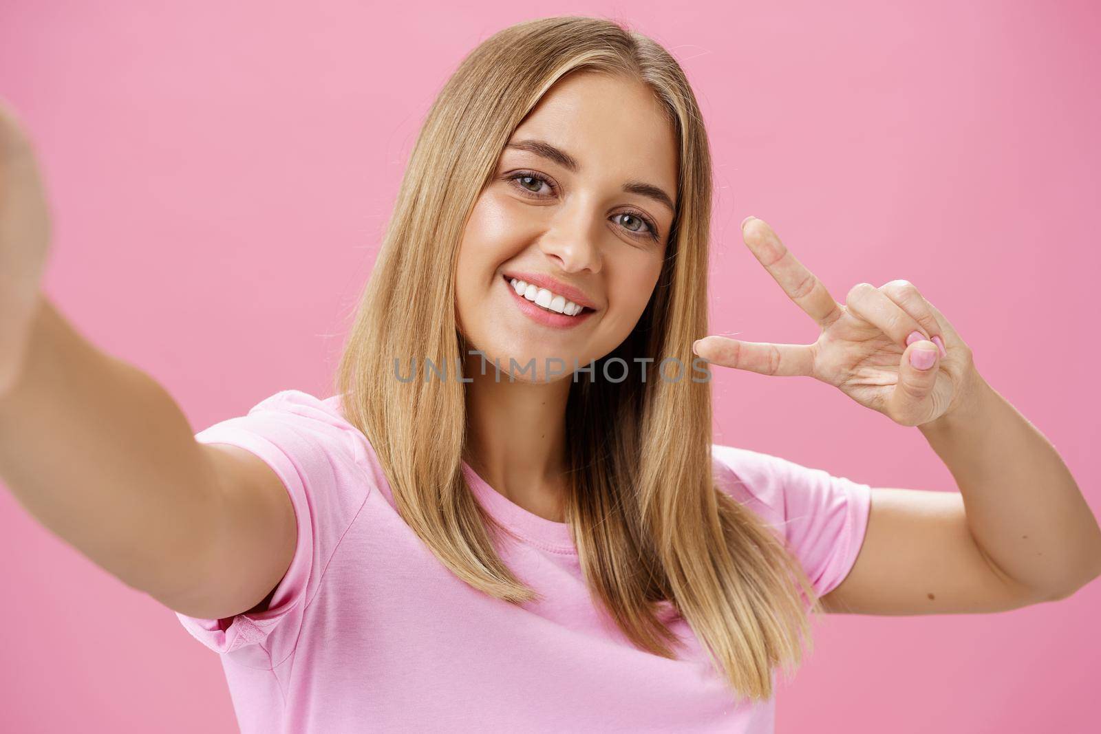 Charming friendly-looking kind young female student with fair hair and chubby face pulling hand towards to hold camera showing peace gesture and smiling broadly taking selfie over pink wall by Benzoix