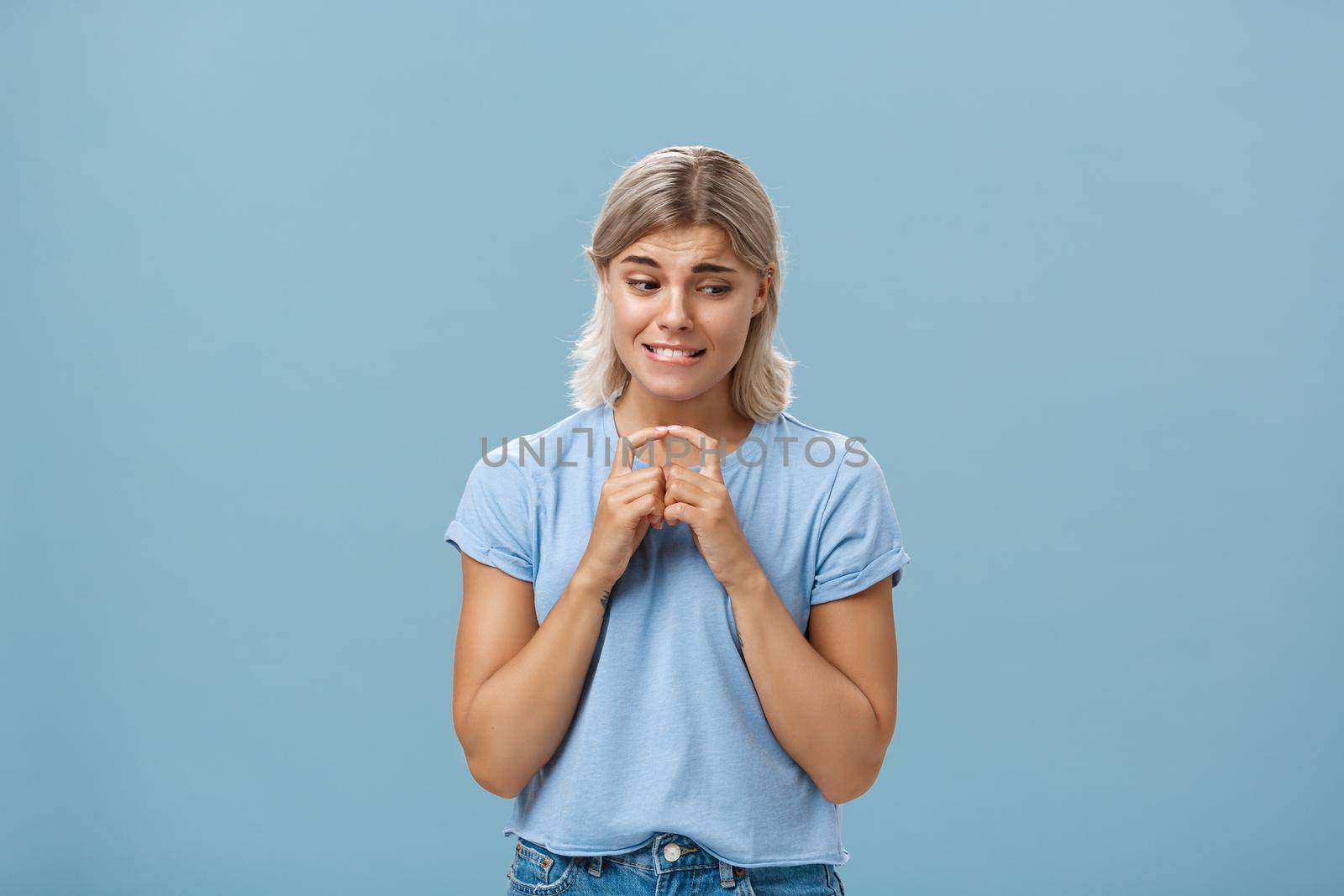 Studio shot of insecure and timid silly girlfriend with blond hair clenching teeth looking shy down and steepling index fingers unconfident trying ask out boy she likes over blue background by Benzoix
