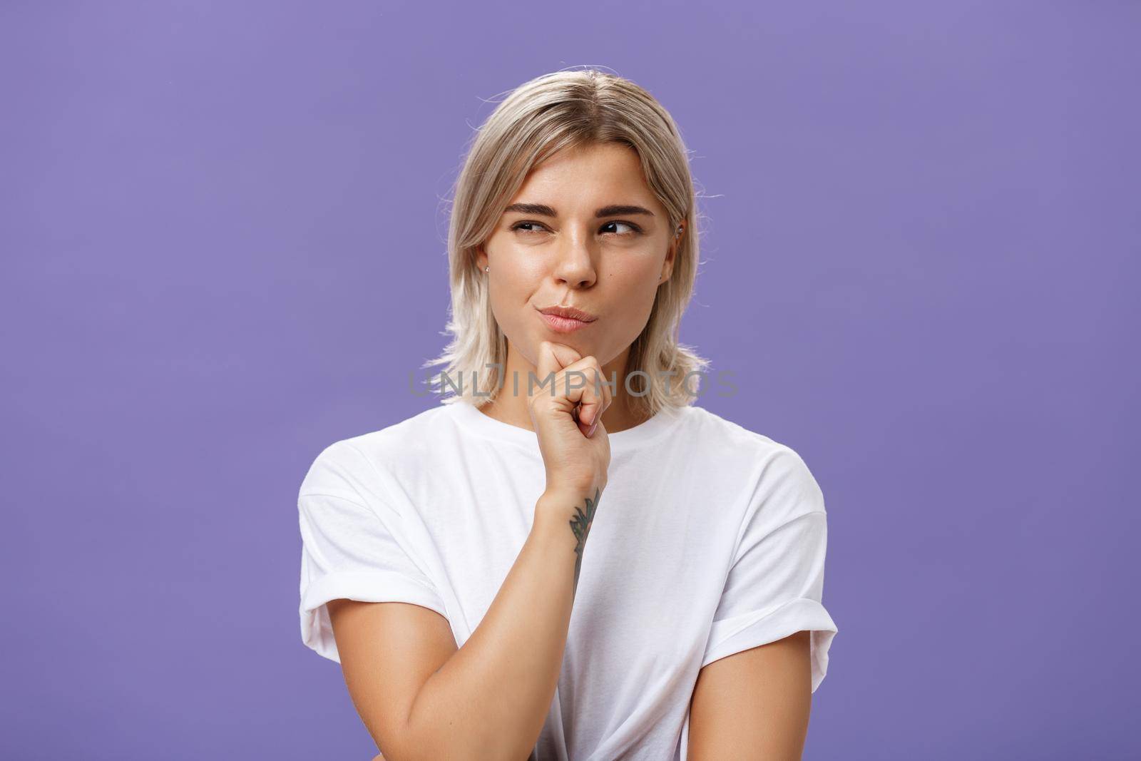 Waist-up shot of smart creative young ambitious woman with blond haircut and tanned skin squinting and smirking gazing left while holding hand on chin thinking making up plan or decision. Copy space