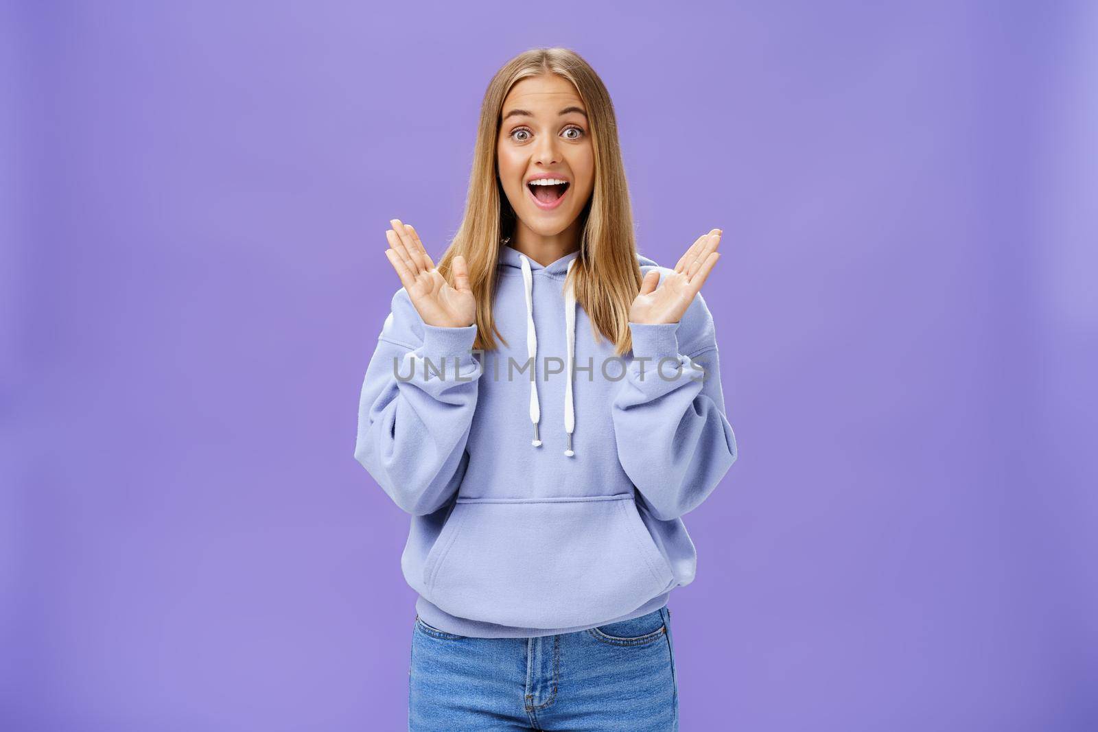 Portrait of amused excited and entertained happy charming woman with fair hair in trendy hoodie clasping hands from delight and surprise smiling broadly joyfully posing against purple background. Lifestyle.