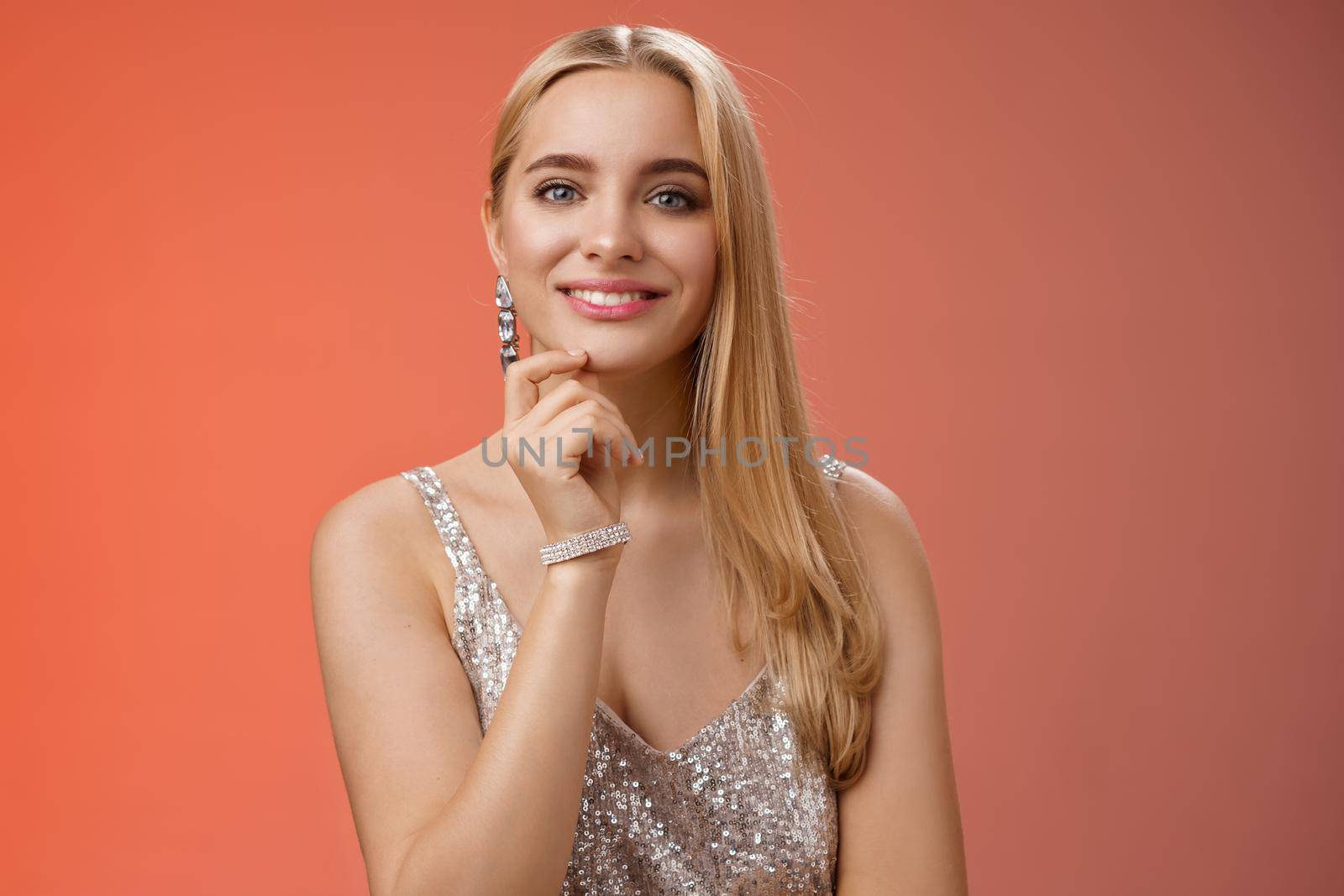 Waist-up shot elegant good-looking wealthy young blond woman in brilliant accessorize silver glittering stylish dress touch chin have clever interesting idea smiling curiously, red background.