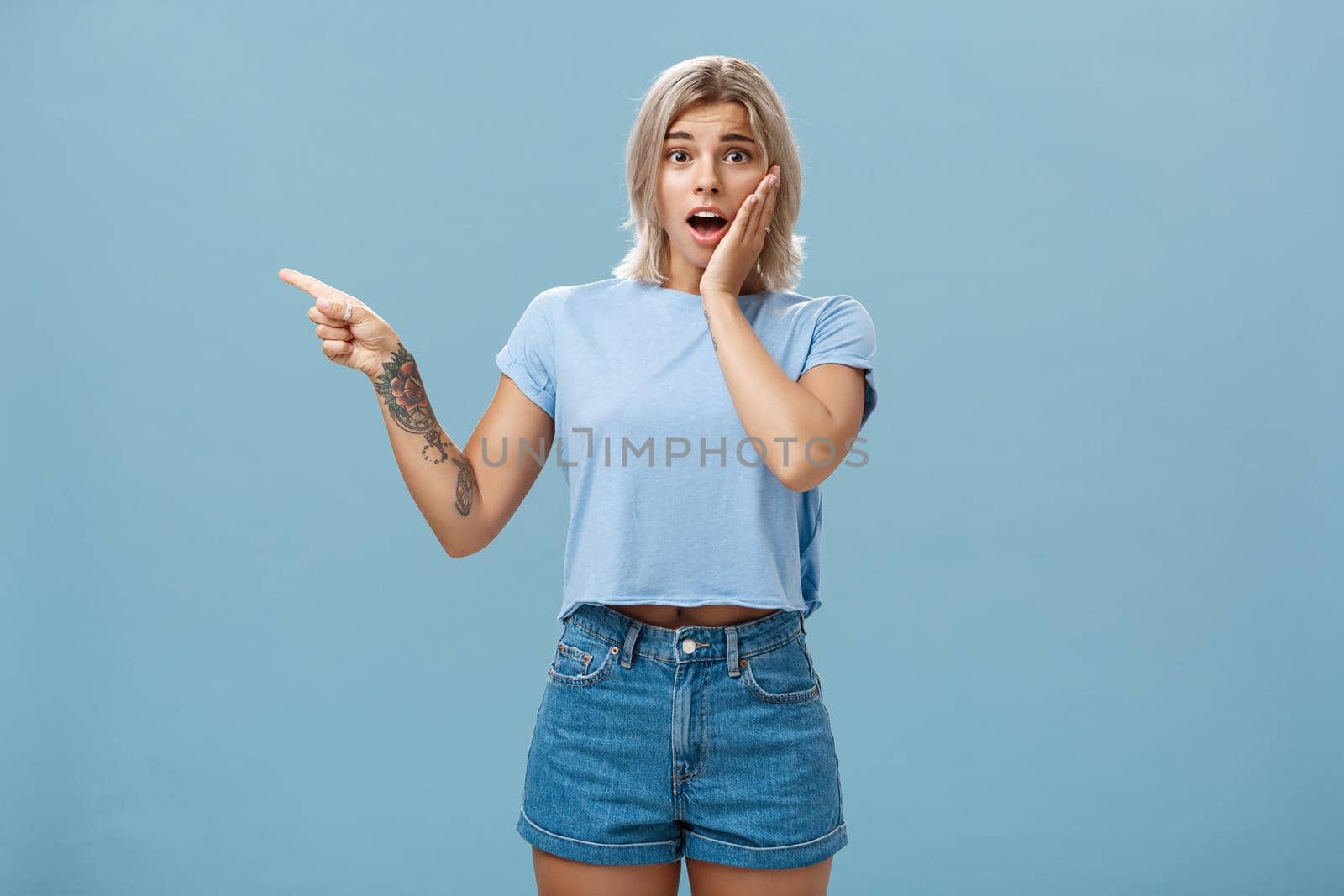 Lifestyle. Waist-up shot of worried surprised emotive blonde woman in t-shirt saying gosh opening mouth from shock holding palm on cheek looking with empathy while pointing left with tattooed arm over blue wall.