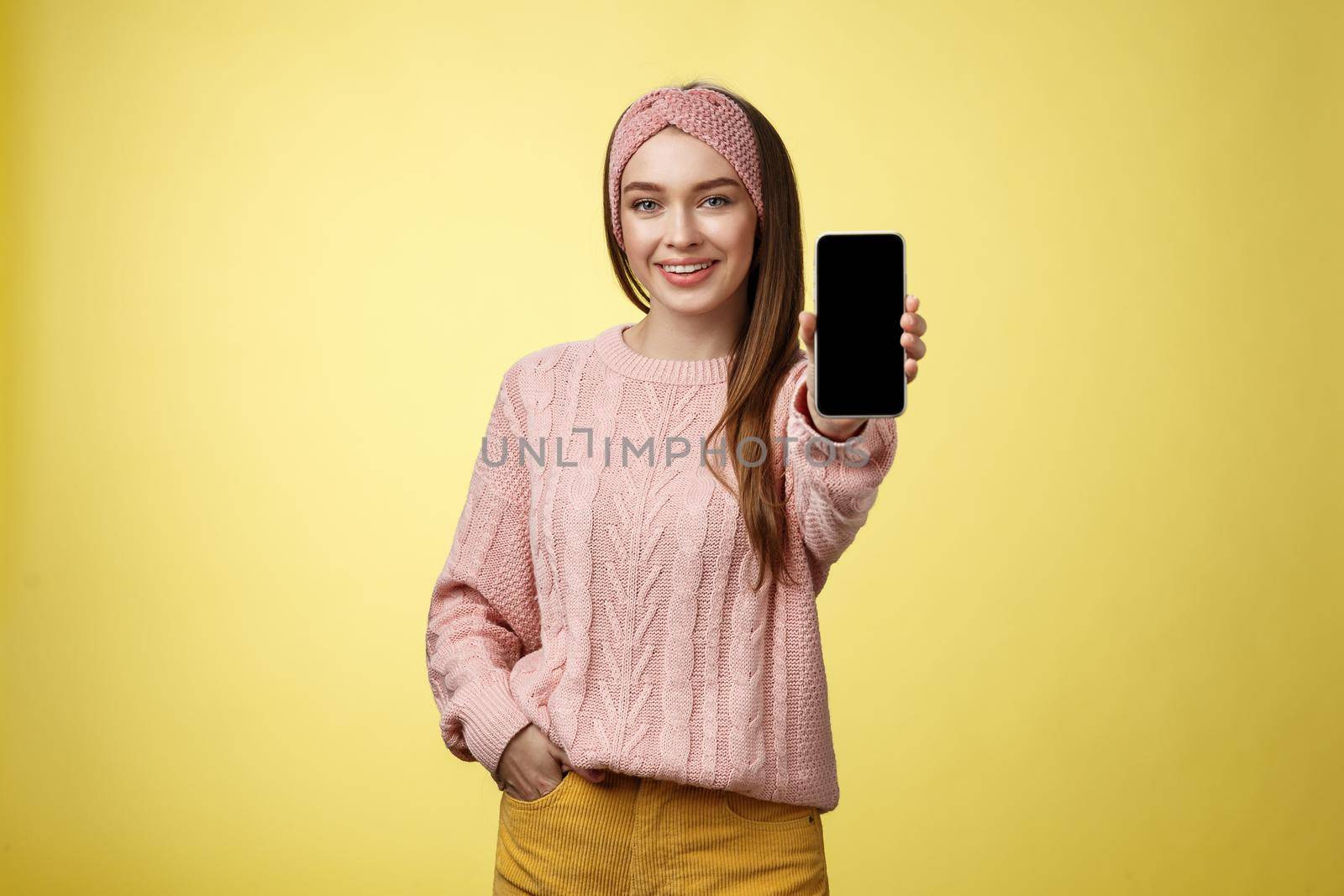 Best smartphone here you go. Charming outgoing young pretty woman in knitted sweater, headband extending hand with phone showing gadget screen smiling recommending cellphone over yellow wall.