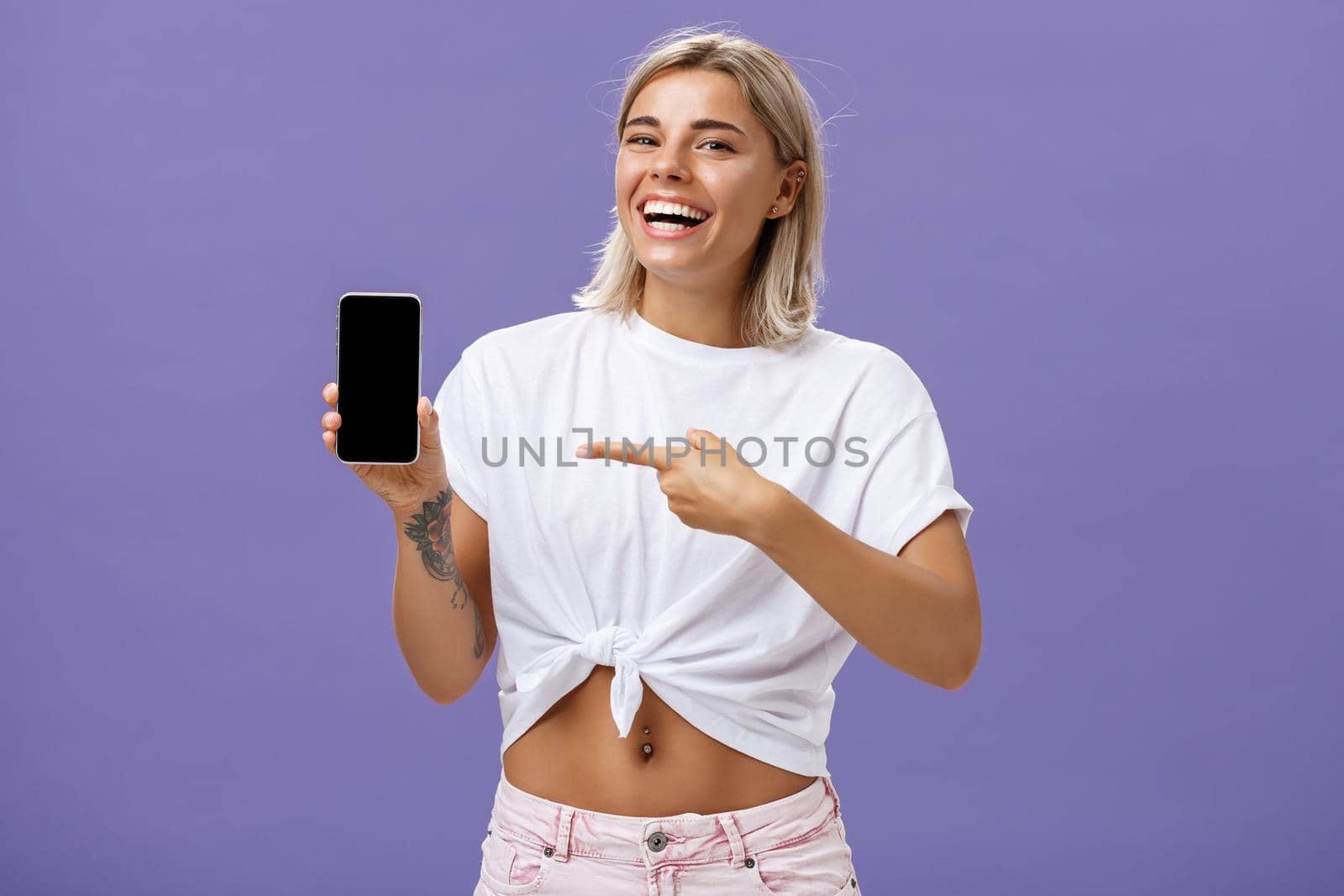 Satisfied happy beautiful tanned woman with blond hairstyle and tattoos holding smartphone showing device screen and pointing at gadget with index finger smiling at camera over purple wall. Technology and advertising concept